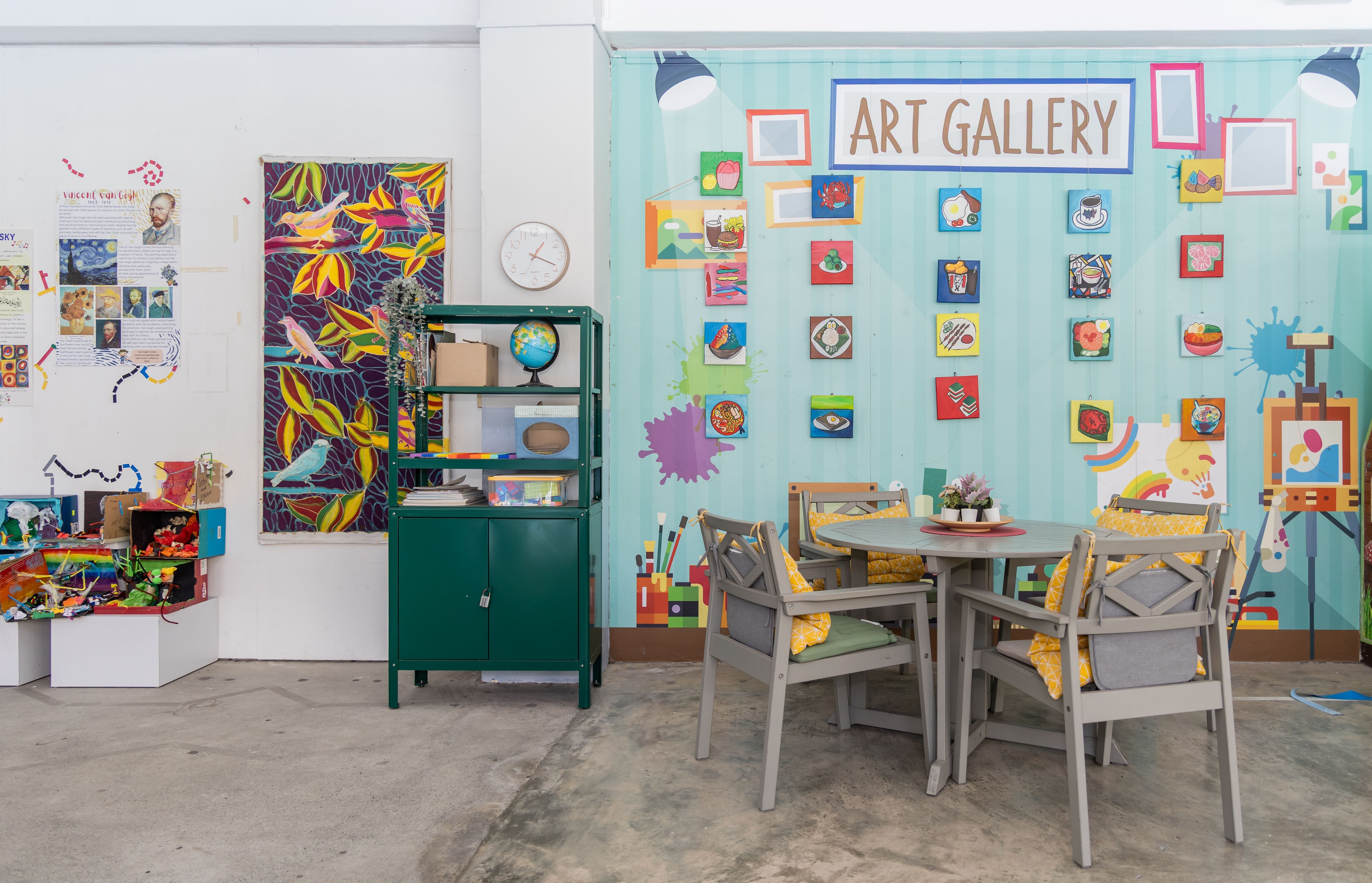 A colorful art gallery room with small paintings on a teal wall, a round table with chairs, and a green metal shelf.