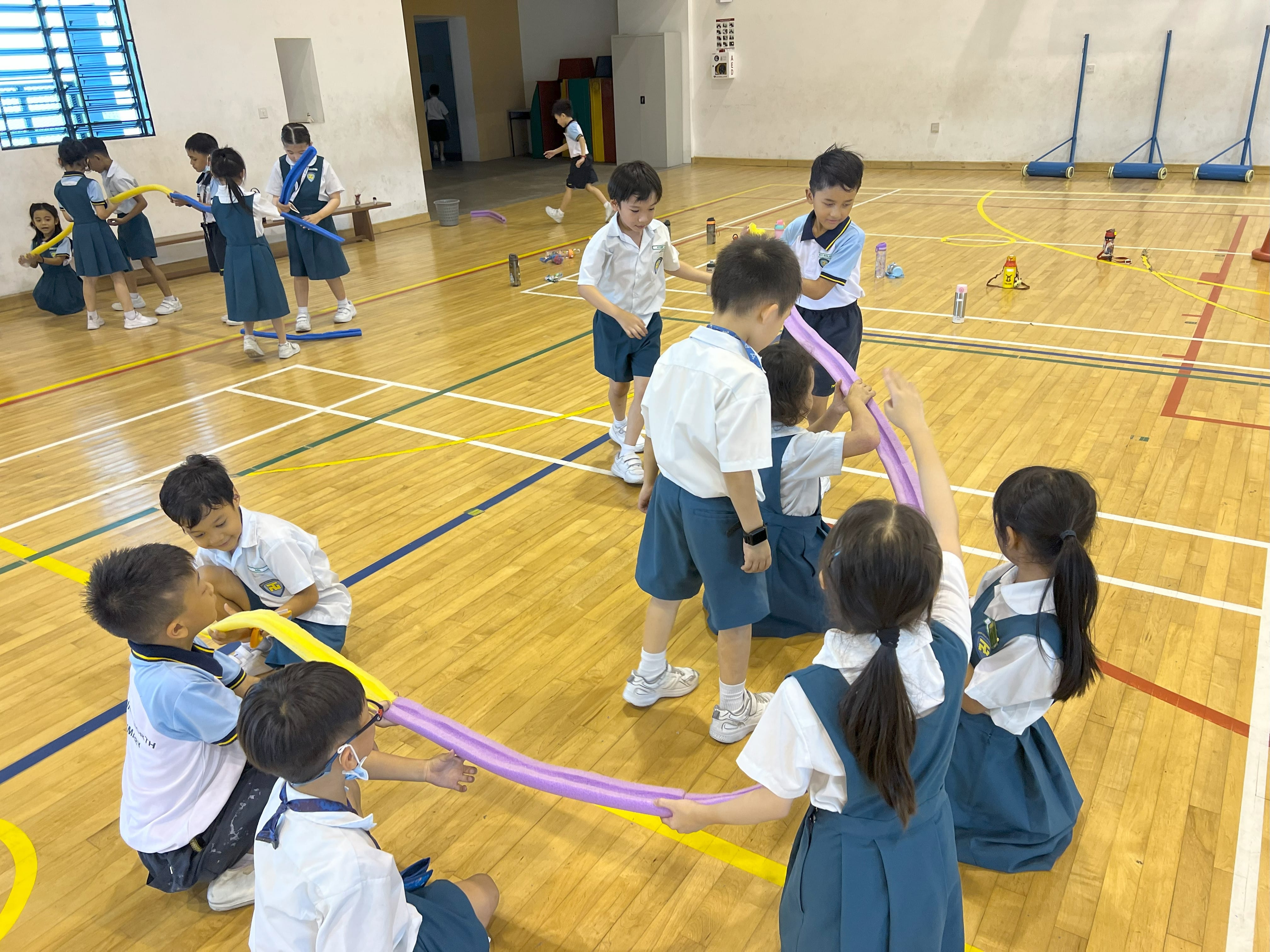 Young school children in uniforms play with colorful foam noodles on a wooden gymnasium floor.