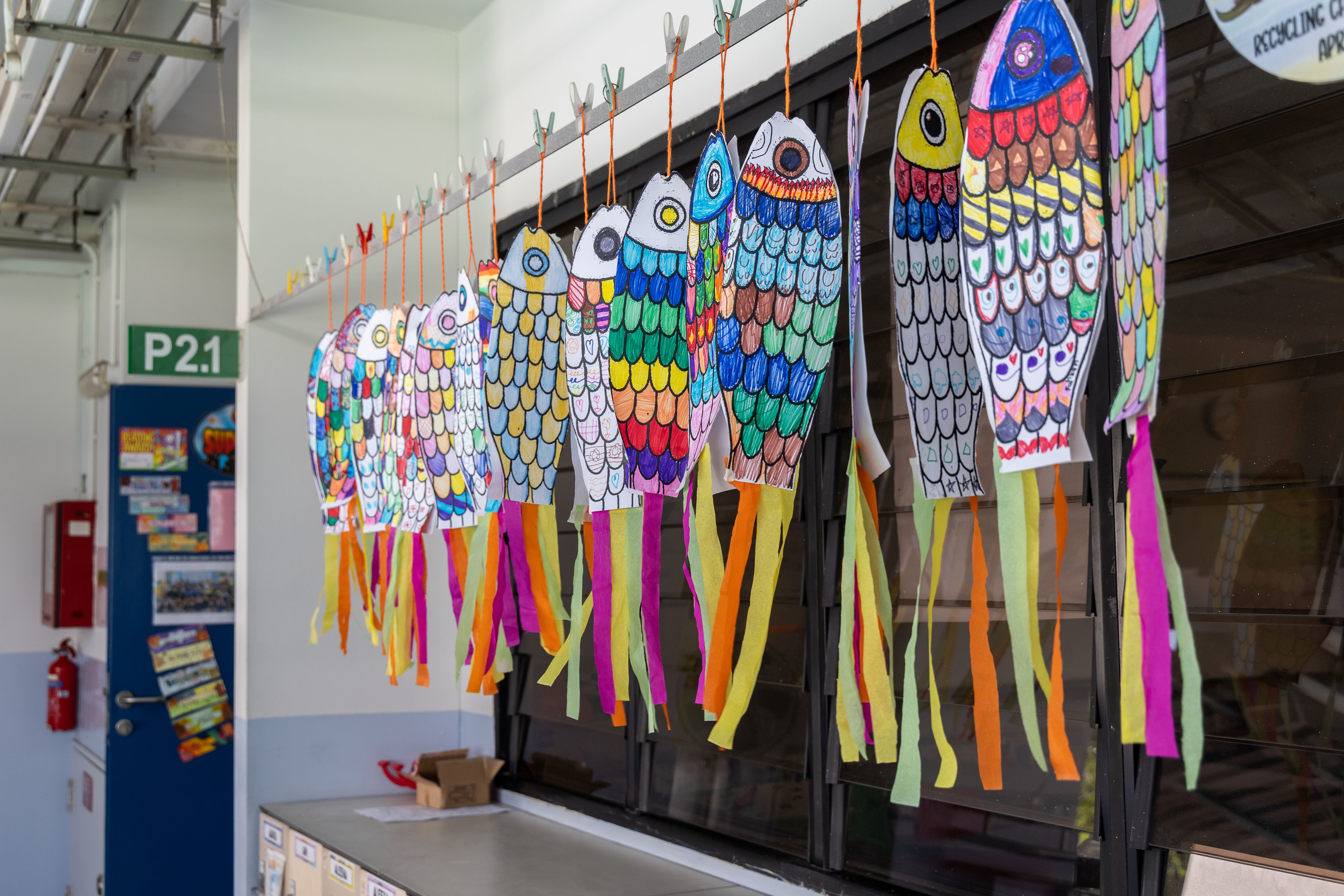 Colorful hand-drawn paper fish with ribbon tails hanging on a line outside a classroom labeled P2.1.
