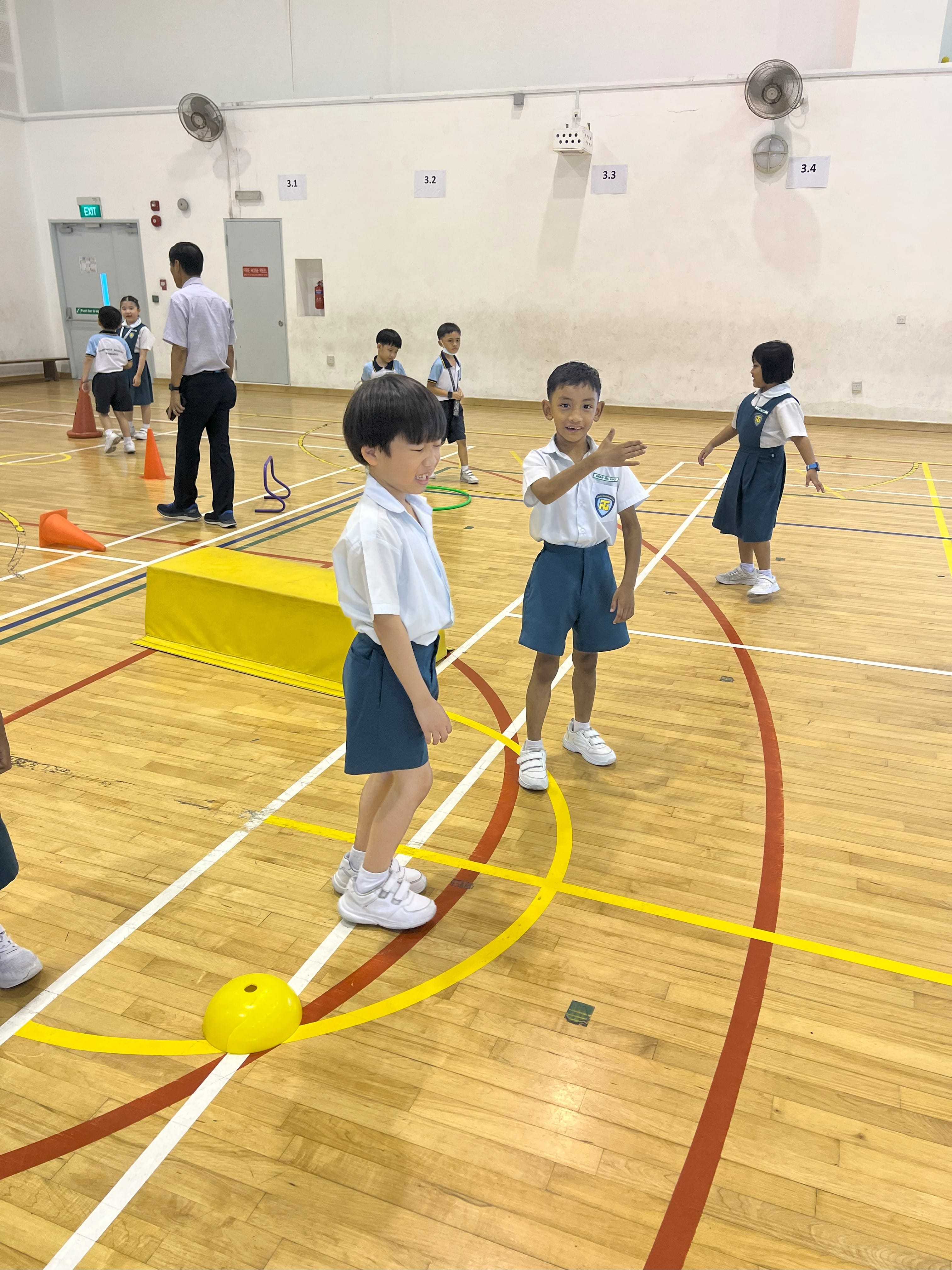 Young children in school uniforms play with a yellow ball on a wooden gymnasium floor with colorful lines.