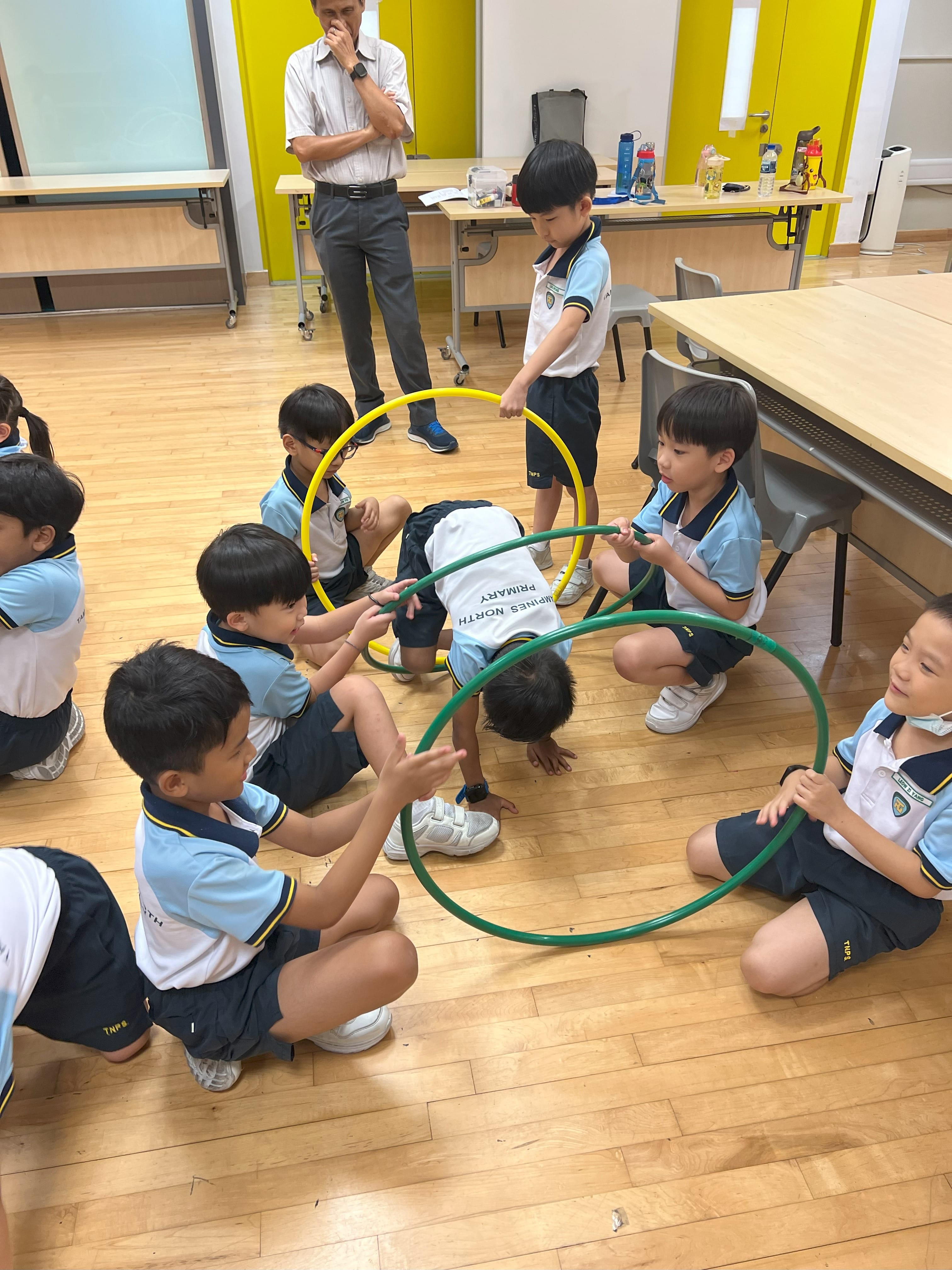 Young students in school uniforms sit on a wooden floor passing green and yellow hoops while an adult watches.