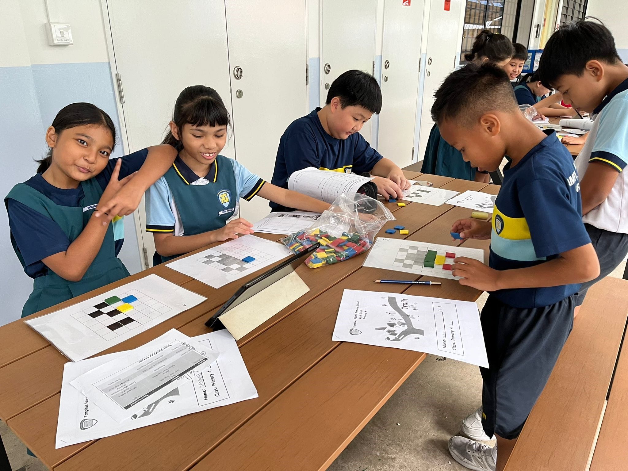 Several primary school children in uniforms work on grid pattern worksheets using colorful tiles at a classroom table.