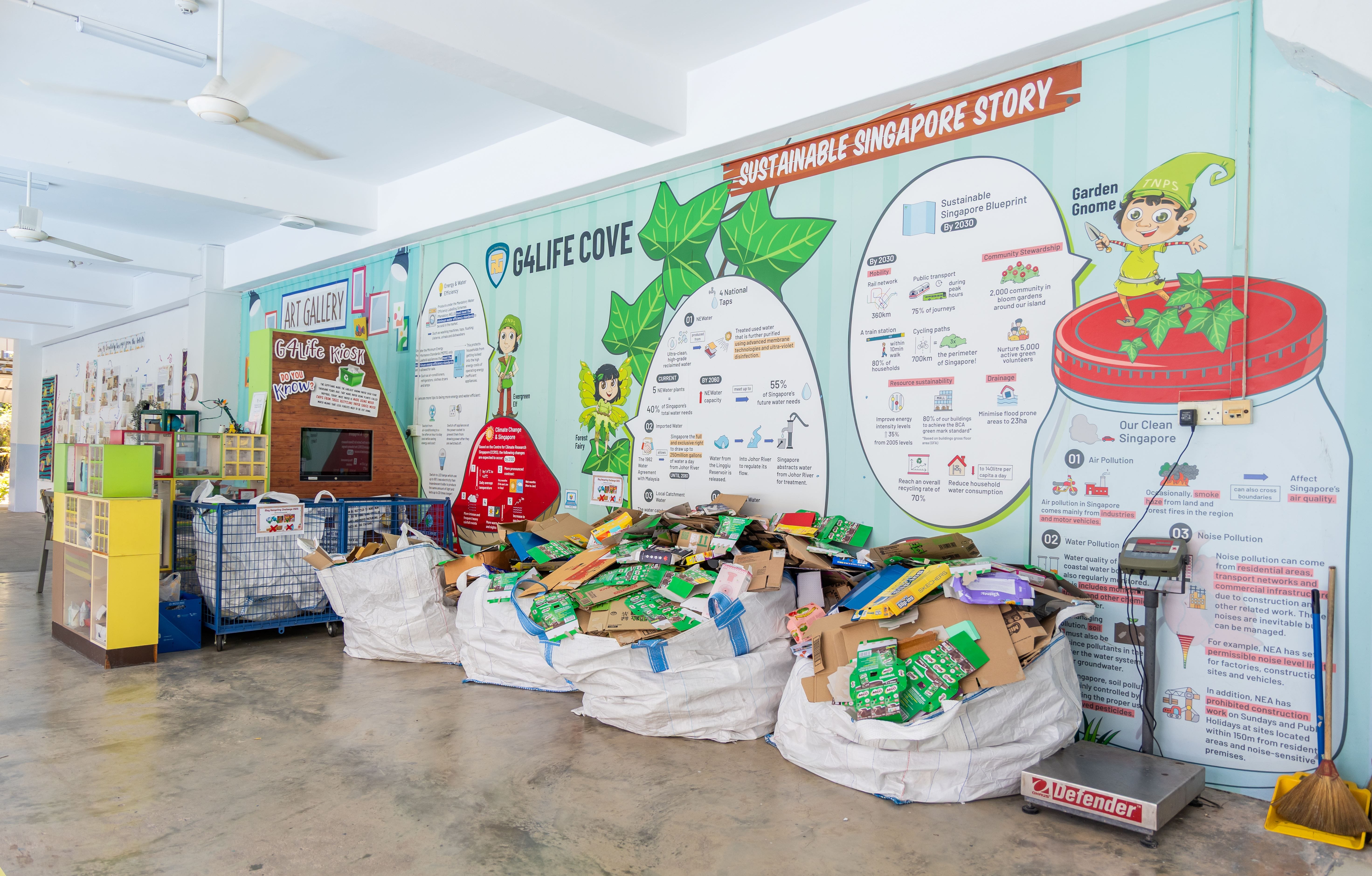 Large bags of recyclable cardboard sit in front of a colorful "Sustainable Singapore Story" educational wall mural.