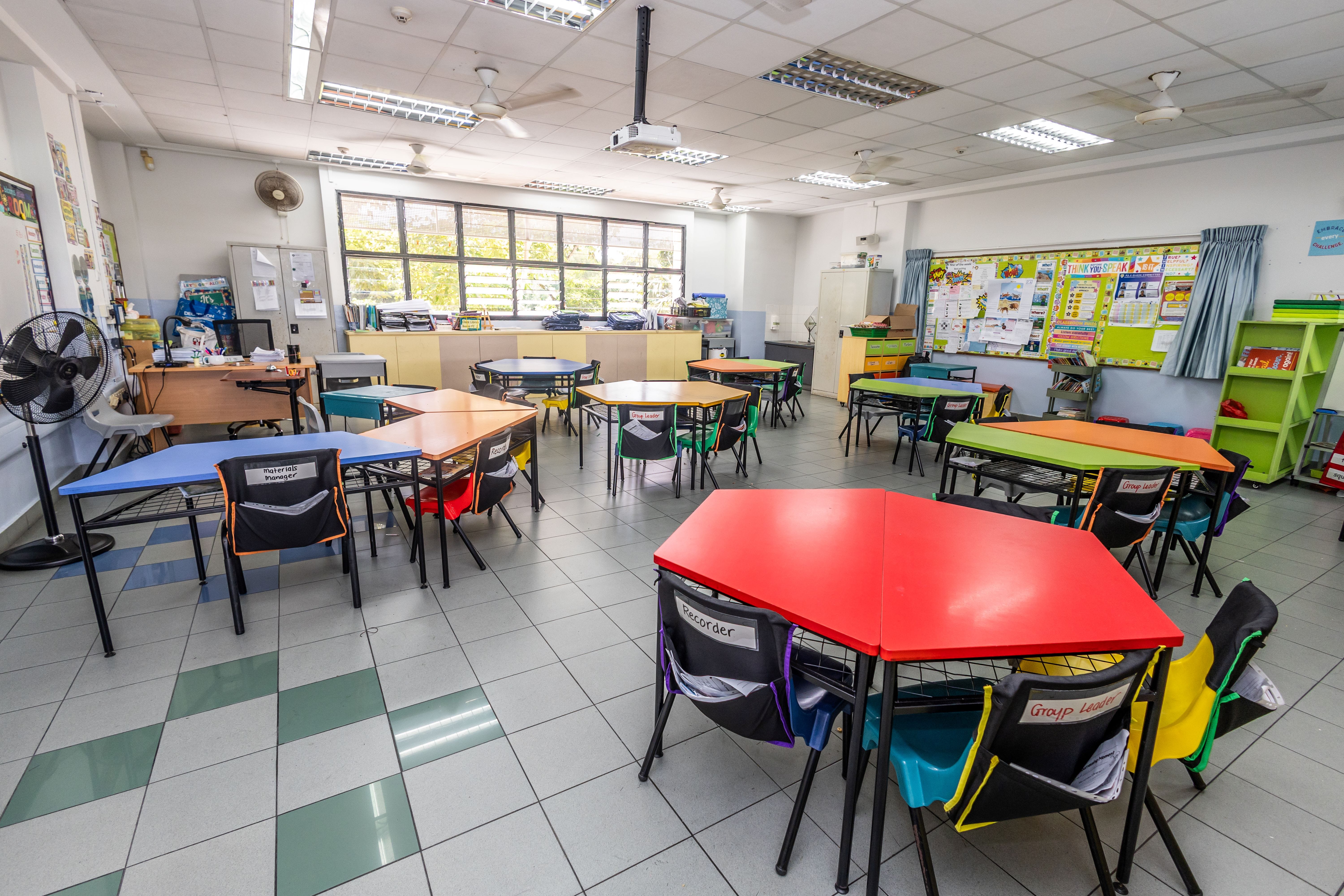 Empty classroom with colorful hexagonal group desks, chairs with pouches, bulletin boards, fans, and a ceiling projector.