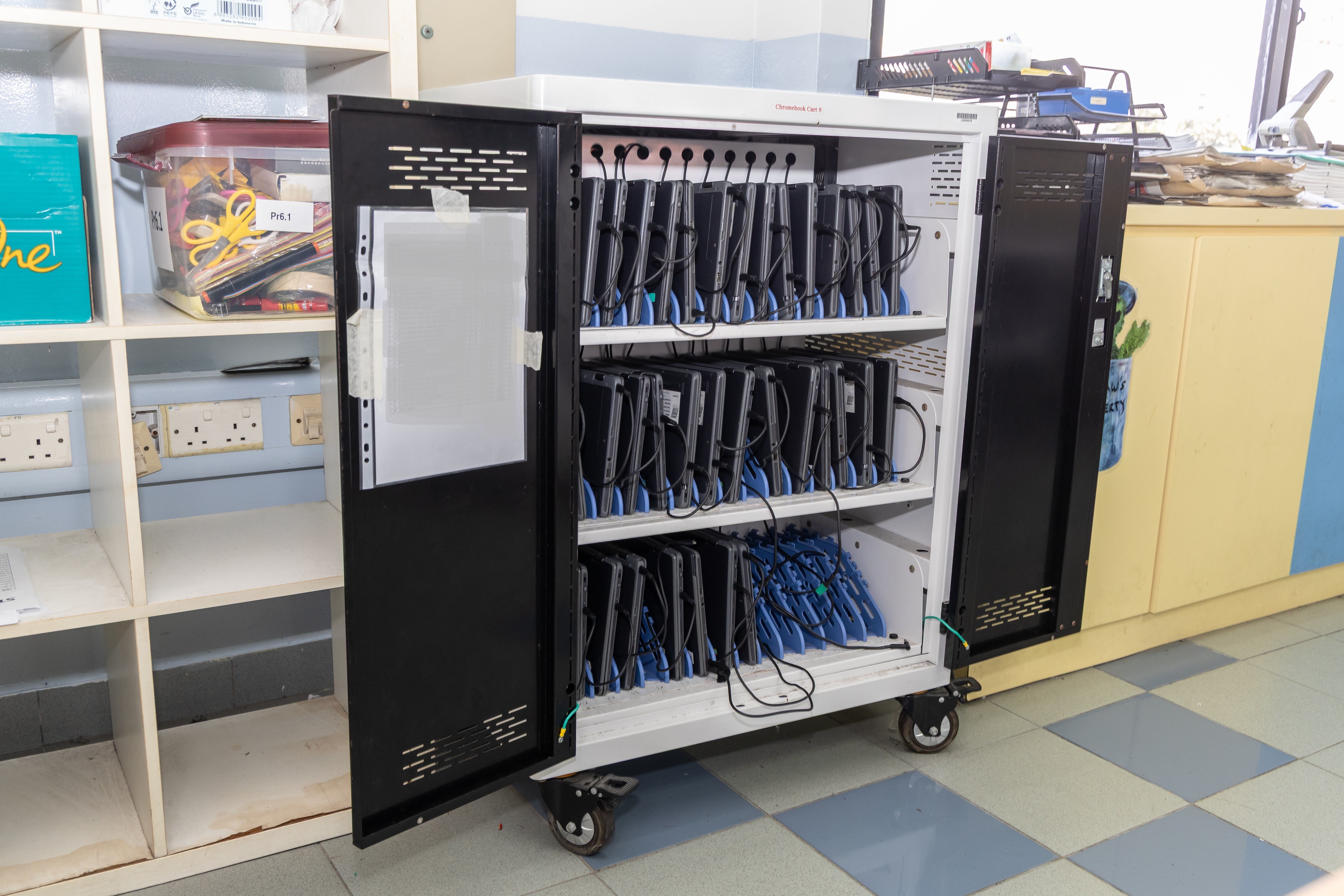 A rolling Chromebook charging cart with open doors revealing three shelves of laptops with cables in a classroom.