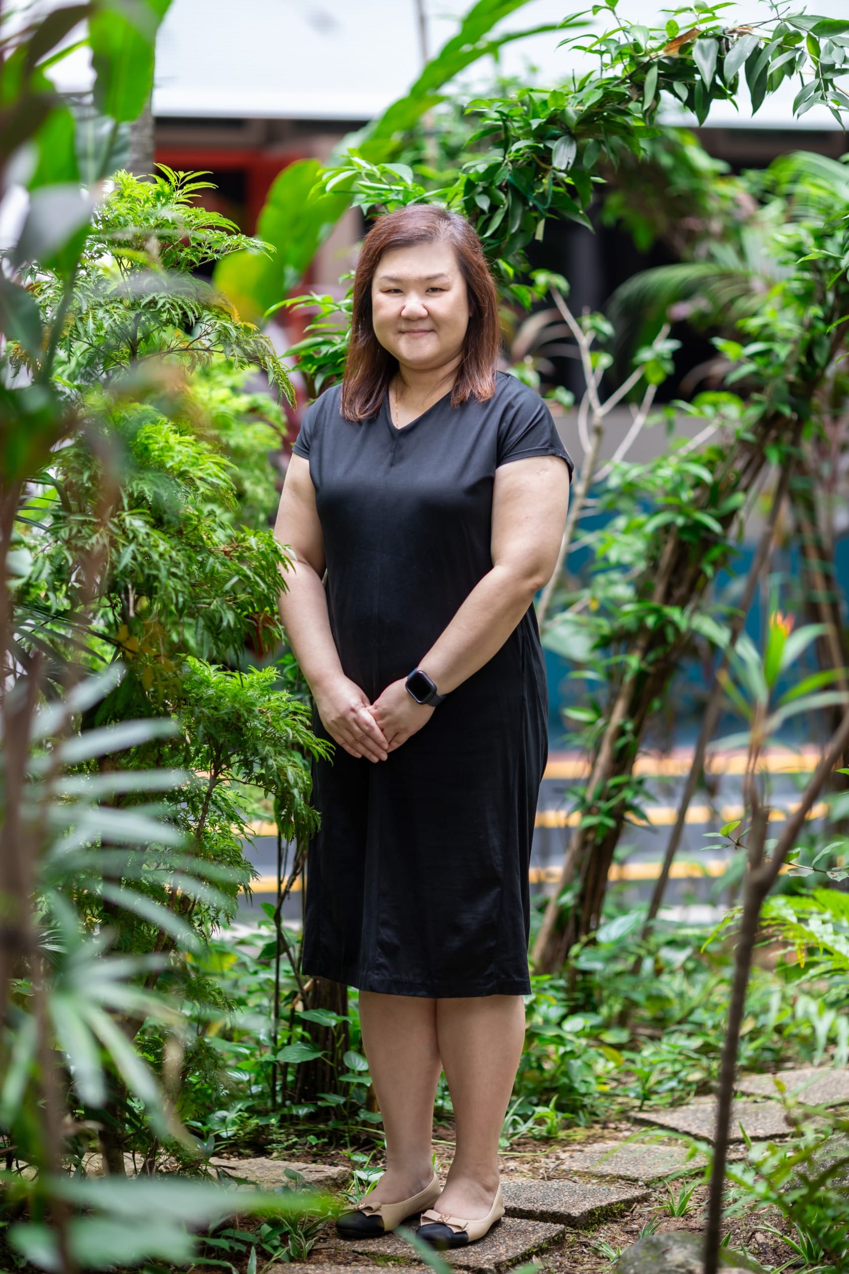 A woman in a black dress stands smiling among green plants on a stone path outdoors.