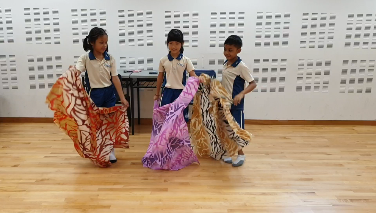 Three children in school uniforms wave large colorful patterned fabric scarves on a wooden floor indoors.