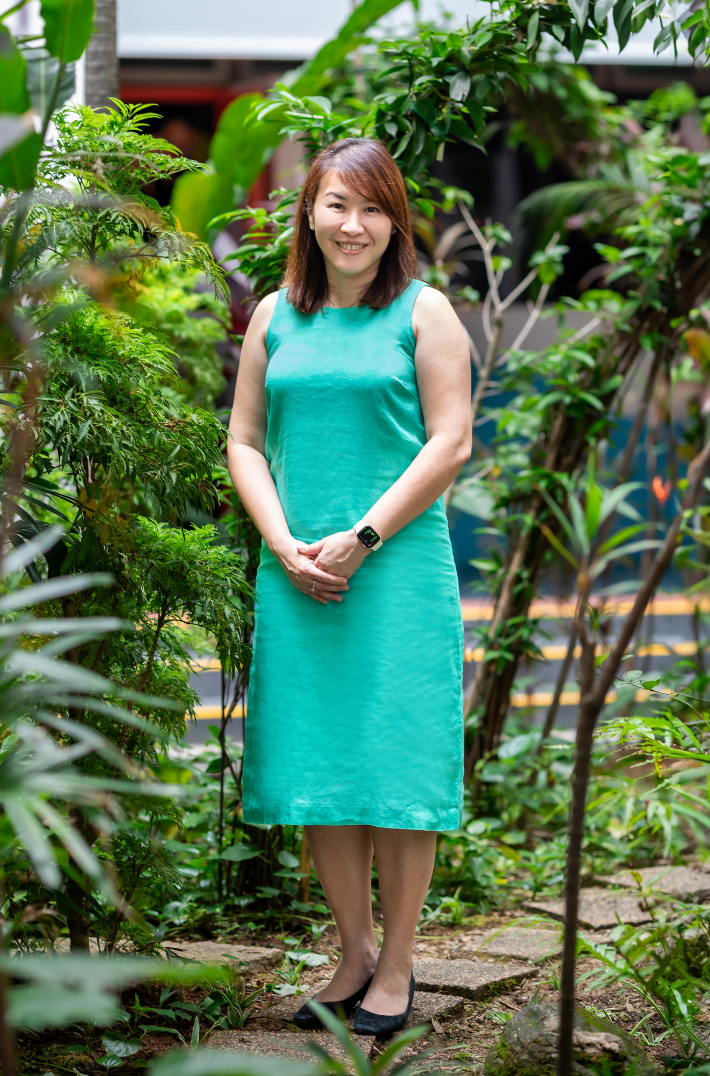A smiling woman in a teal sleeveless dress stands among green plants outdoors, hands clasped, wearing a smartwatch.