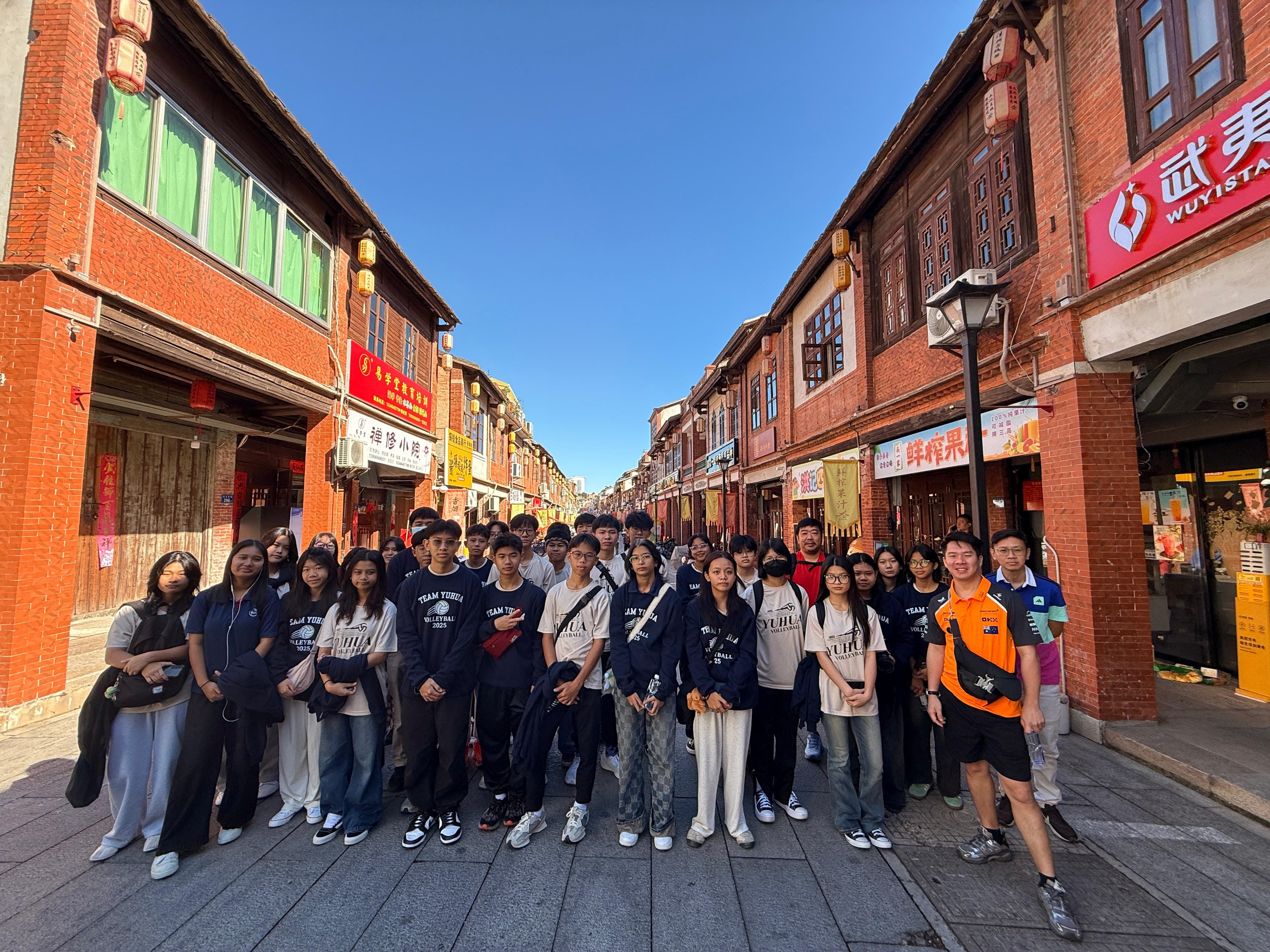 A large group of young people in matching shirts pose on a traditional Chinese street lined with red brick buildings.