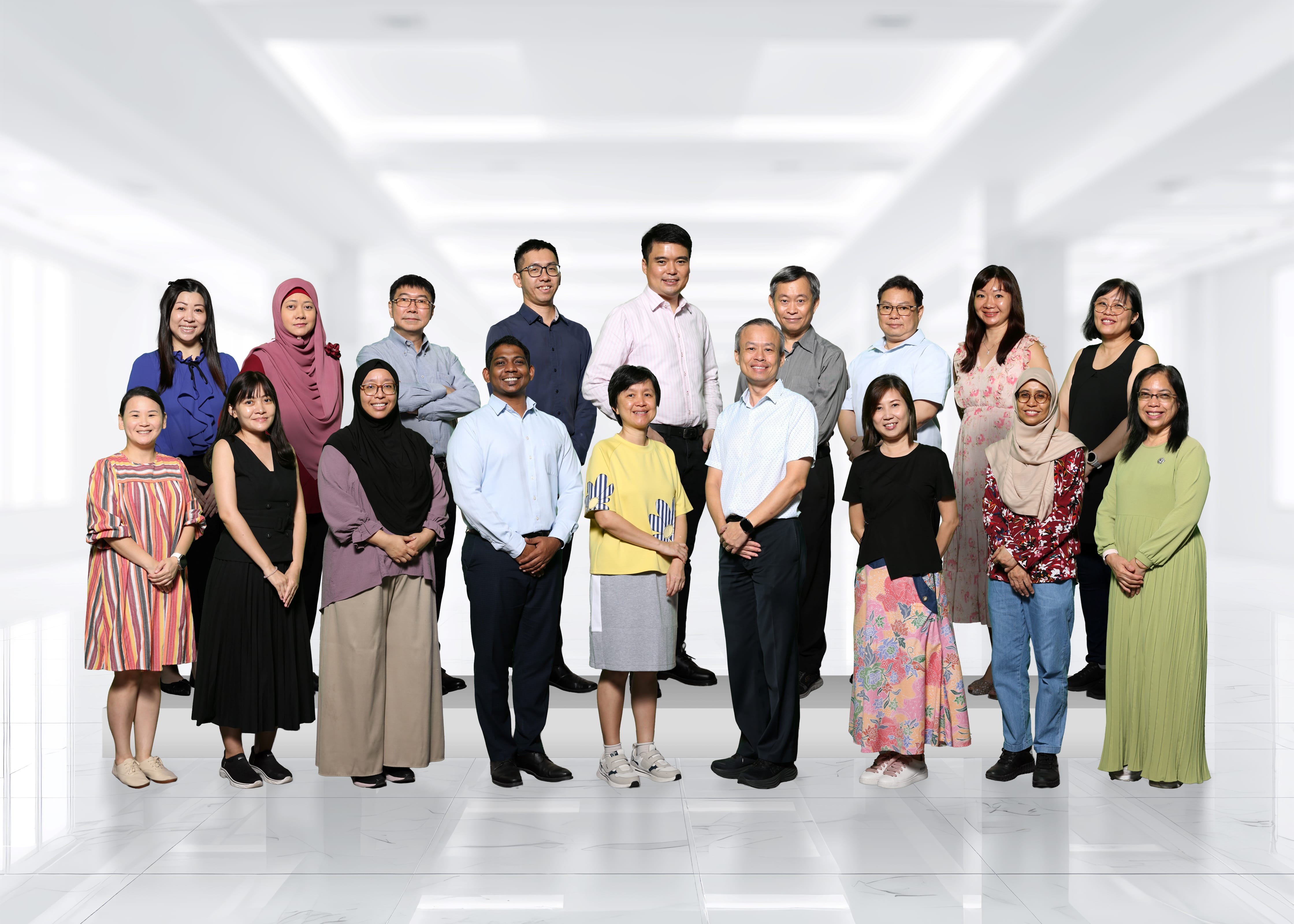 A group of about 18 adults posing together in two rows inside a bright white corridor.