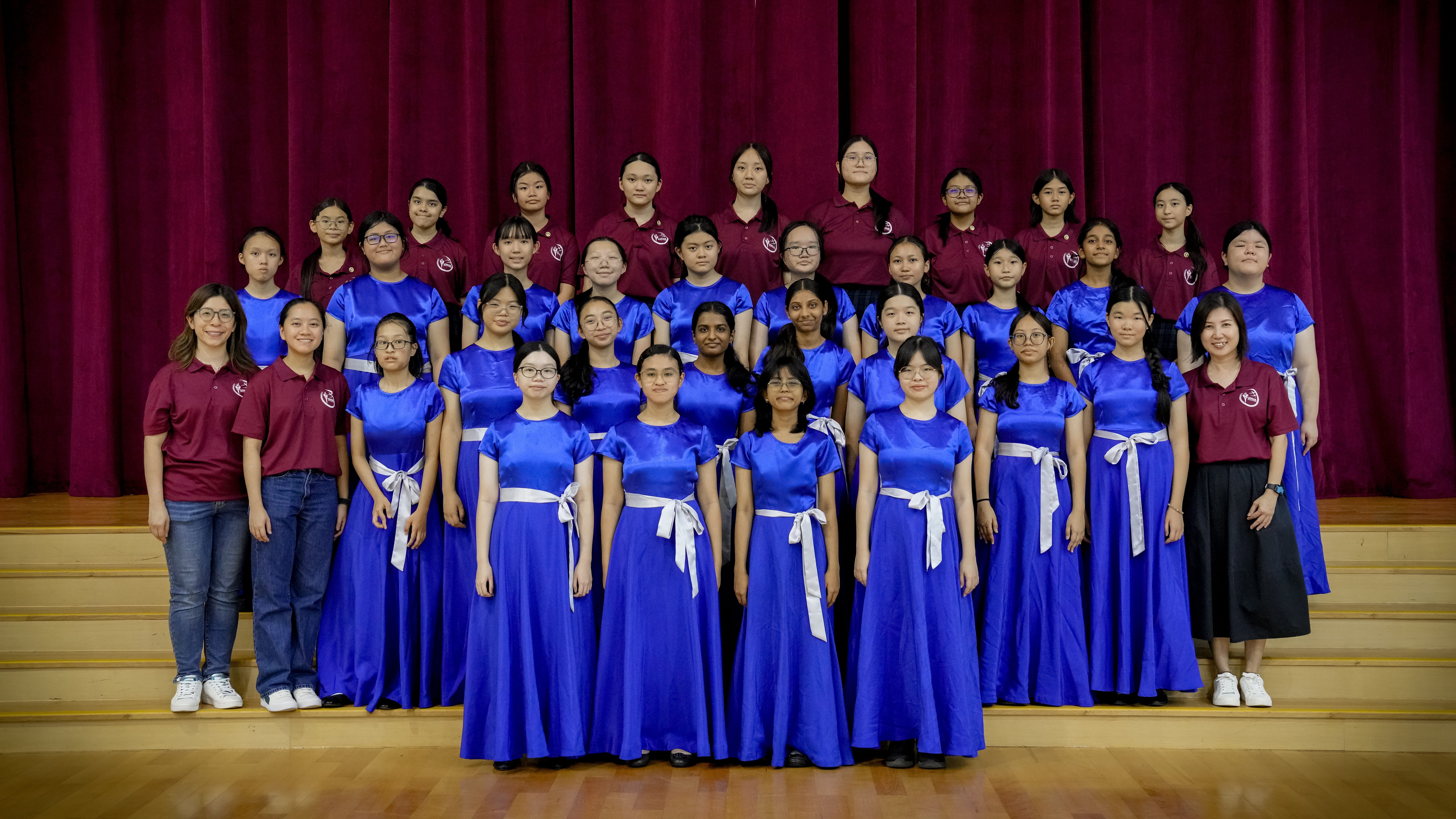 Group photo on stage: teens in blue gowns and maroon polos, with two adult women at the sides.