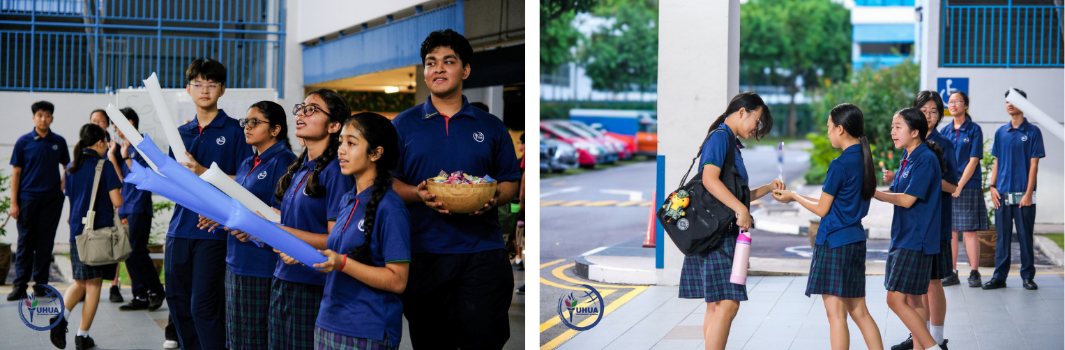 Students in blue uniforms gather outdoors, some holding paper rolls and a bowl of candy, others chatting near a school entrance.