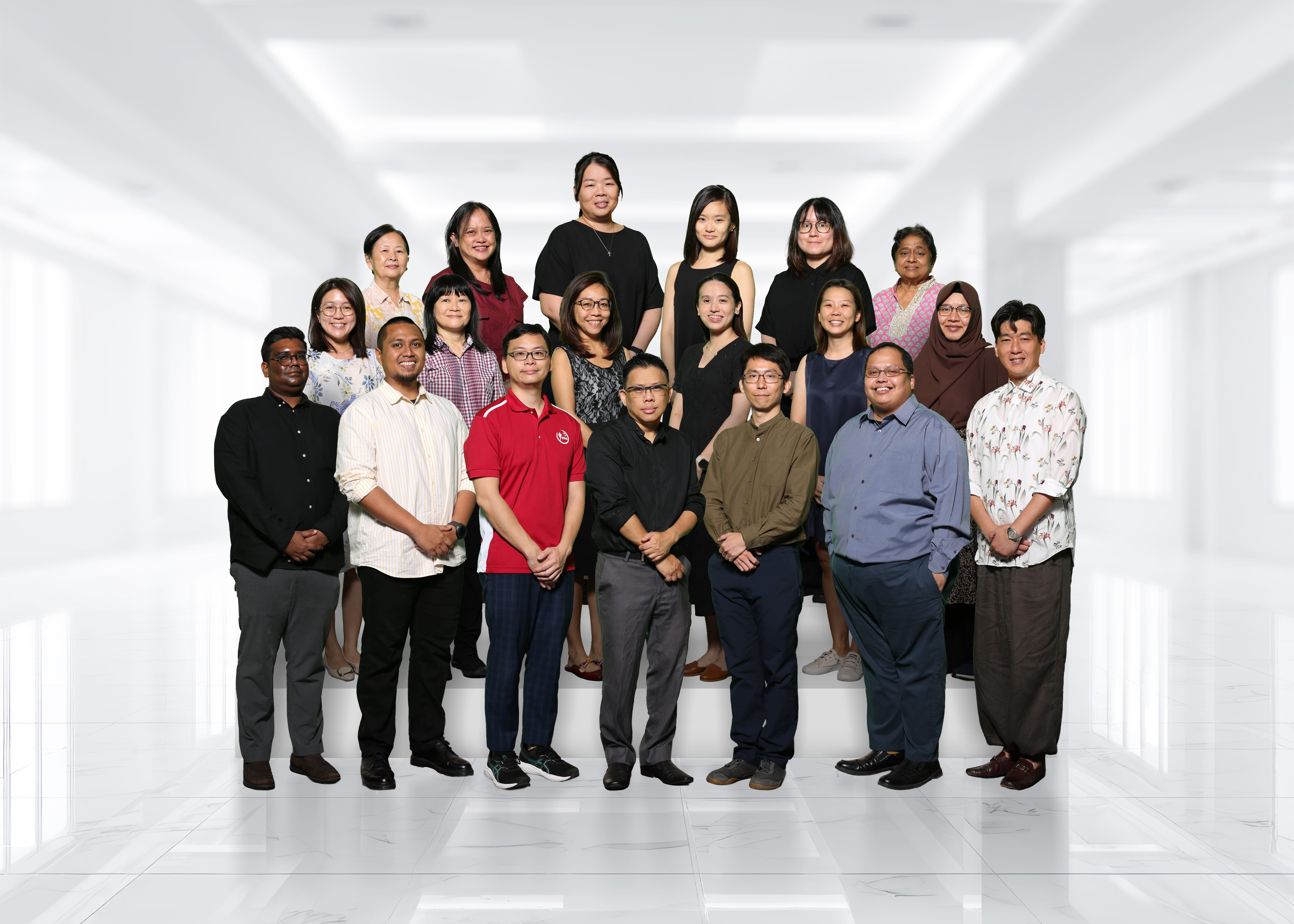 Group portrait of 18 people posing in three rows in a bright white hallway, wearing business casual clothes.