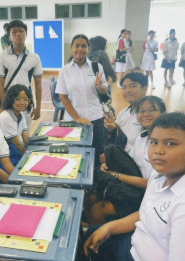 Students in white uniforms gather around a table with folders and devices in a school hall.