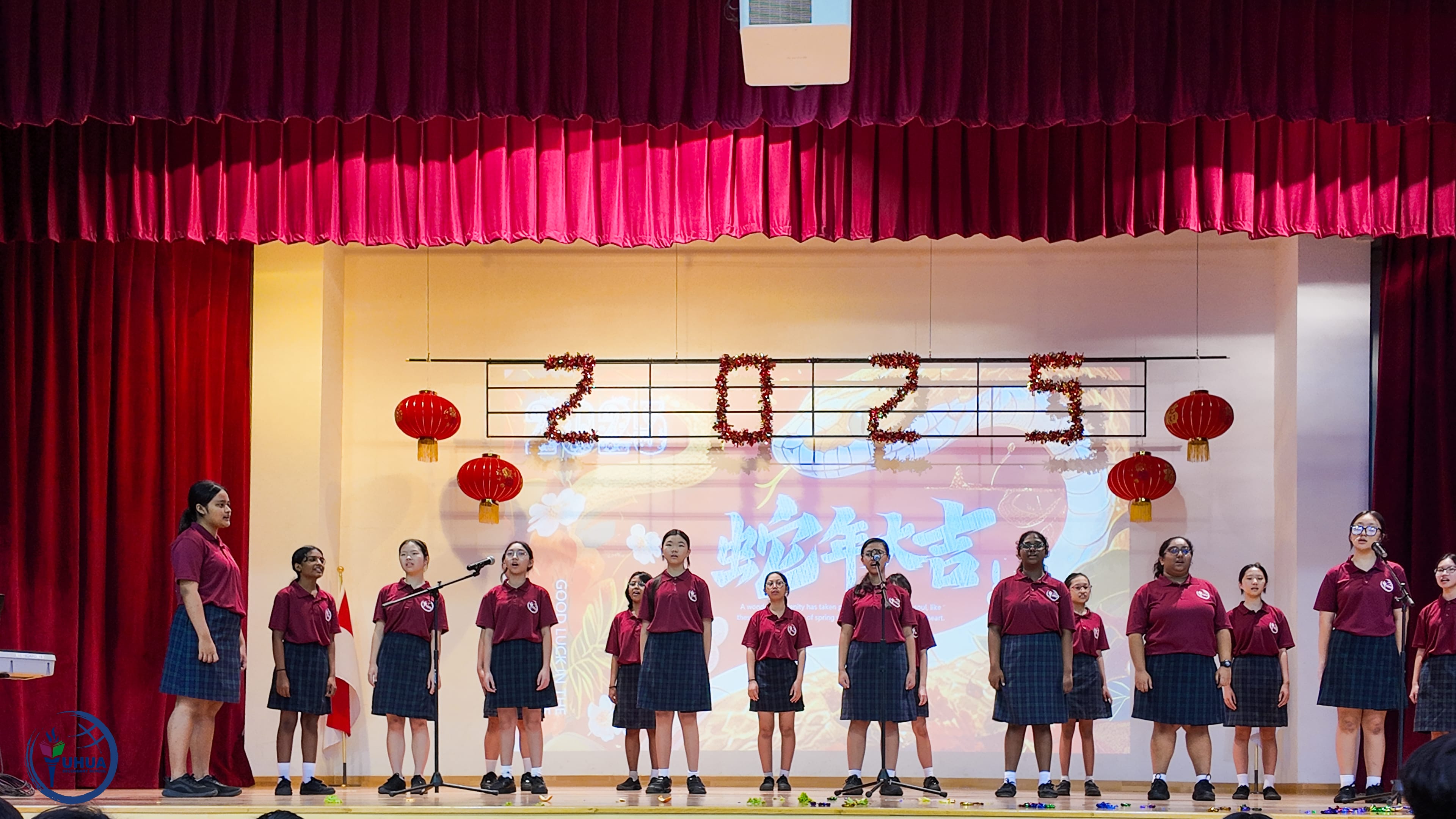 Choir of students in maroon uniforms on stage under 2025 sign with red lanterns and screen backdrop.