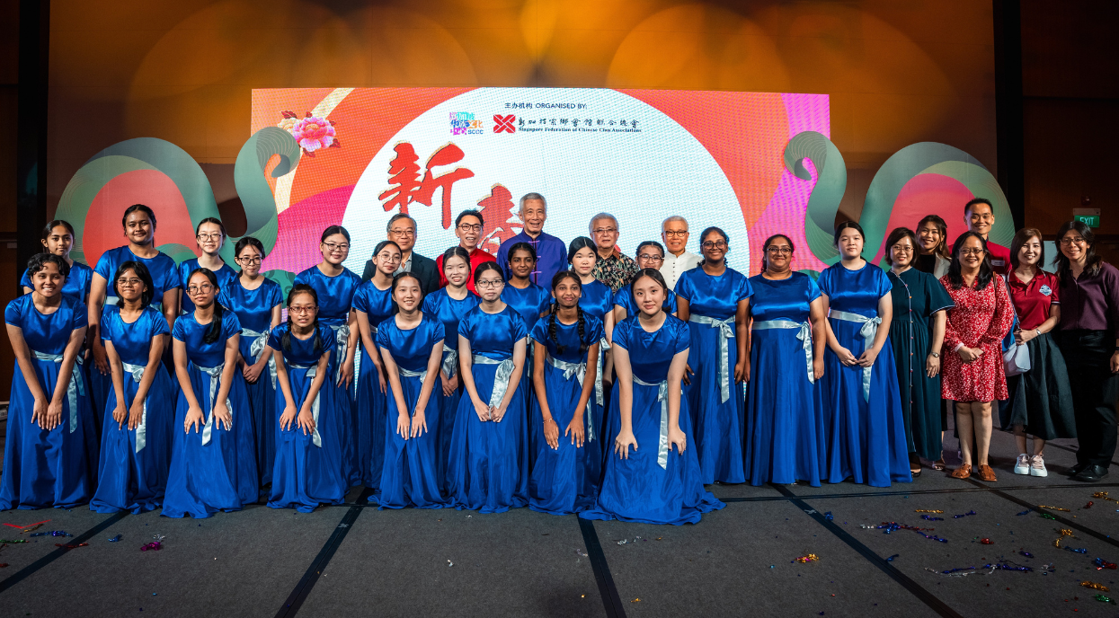 A large group poses in front of a colorful Chinese New Year backdrop, with young performers in blue satin gowns.