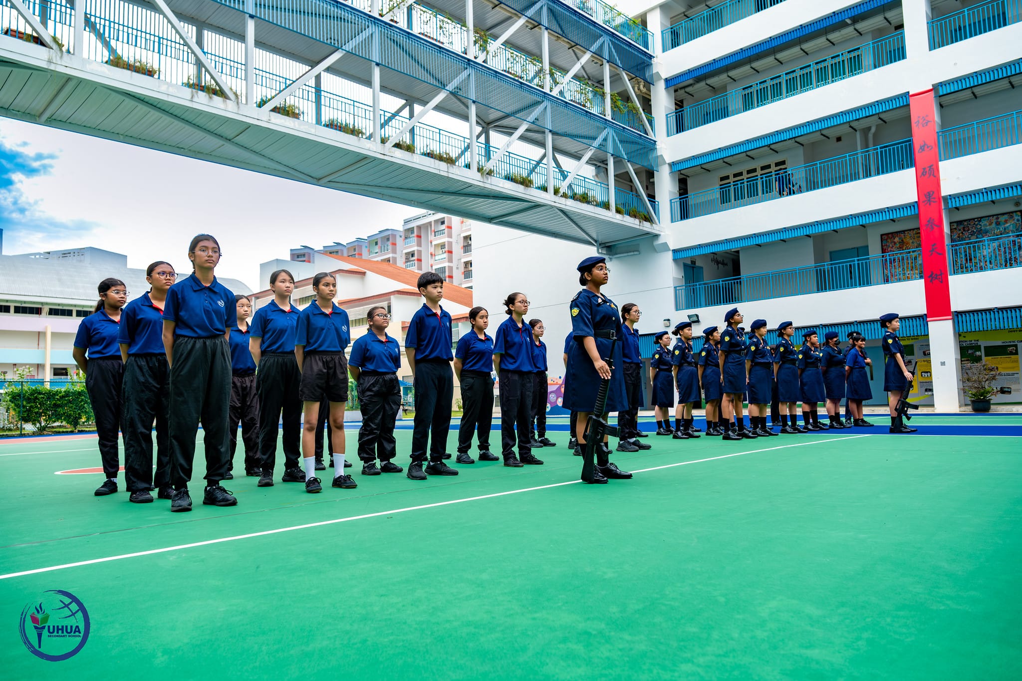 Students in blue uniforms stand in formation on a green school courtyard, with a multi-story school building behind them.
