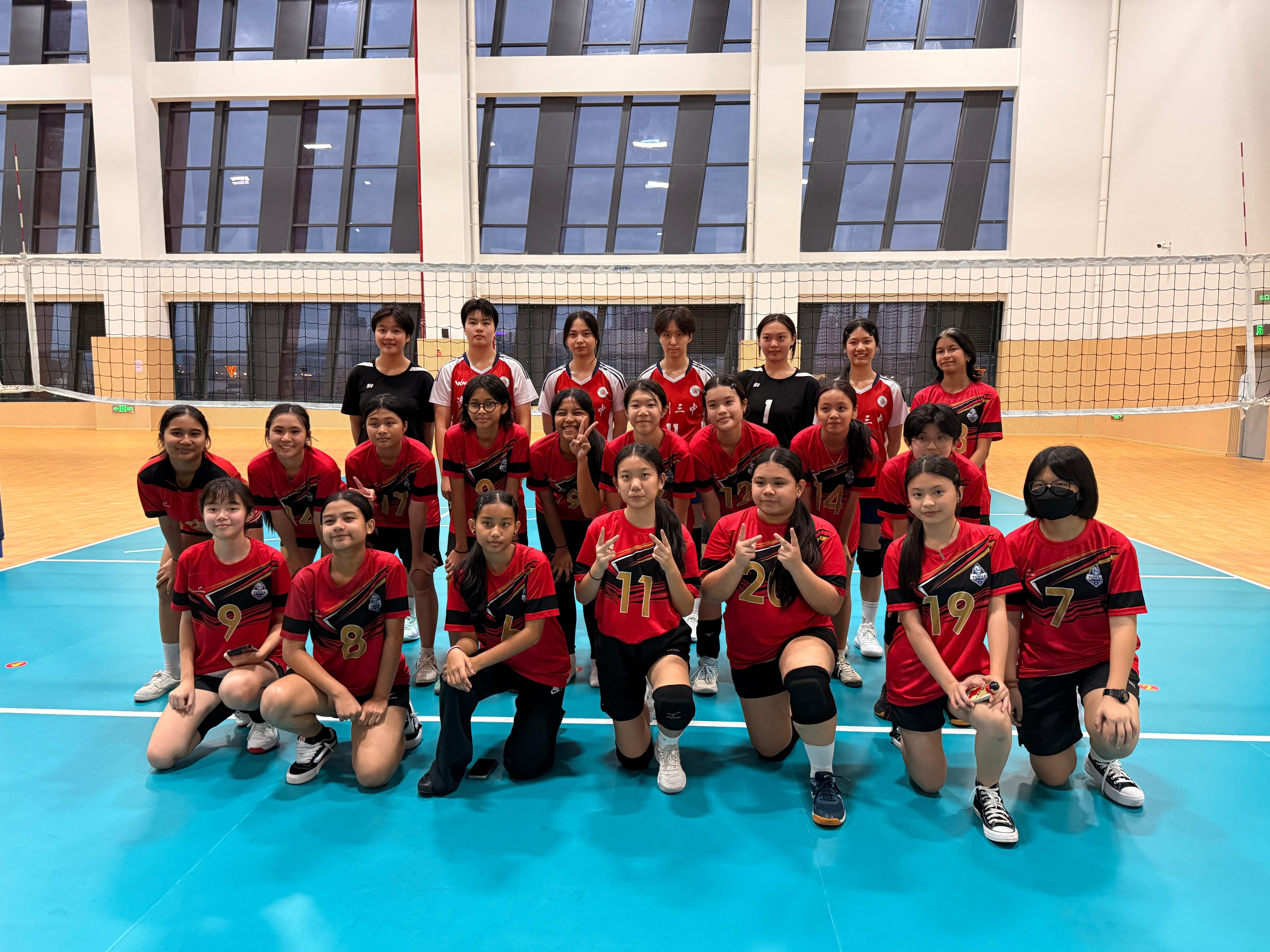 A girls volleyball team in red and black uniforms poses together on an indoor court in front of a net.