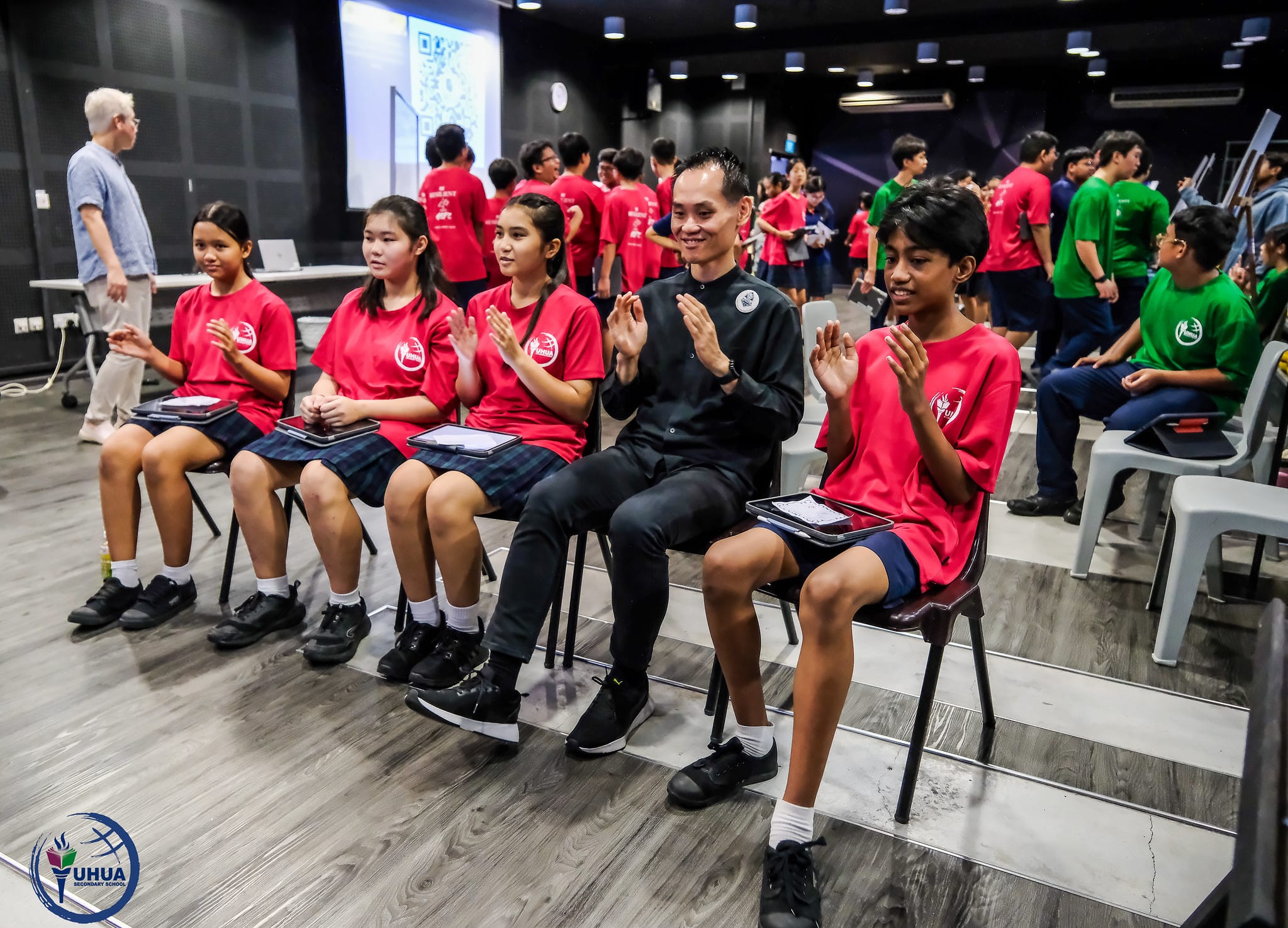 Students in red shirts and a teacher in black sit clapping in a school hall with more students standing behind.