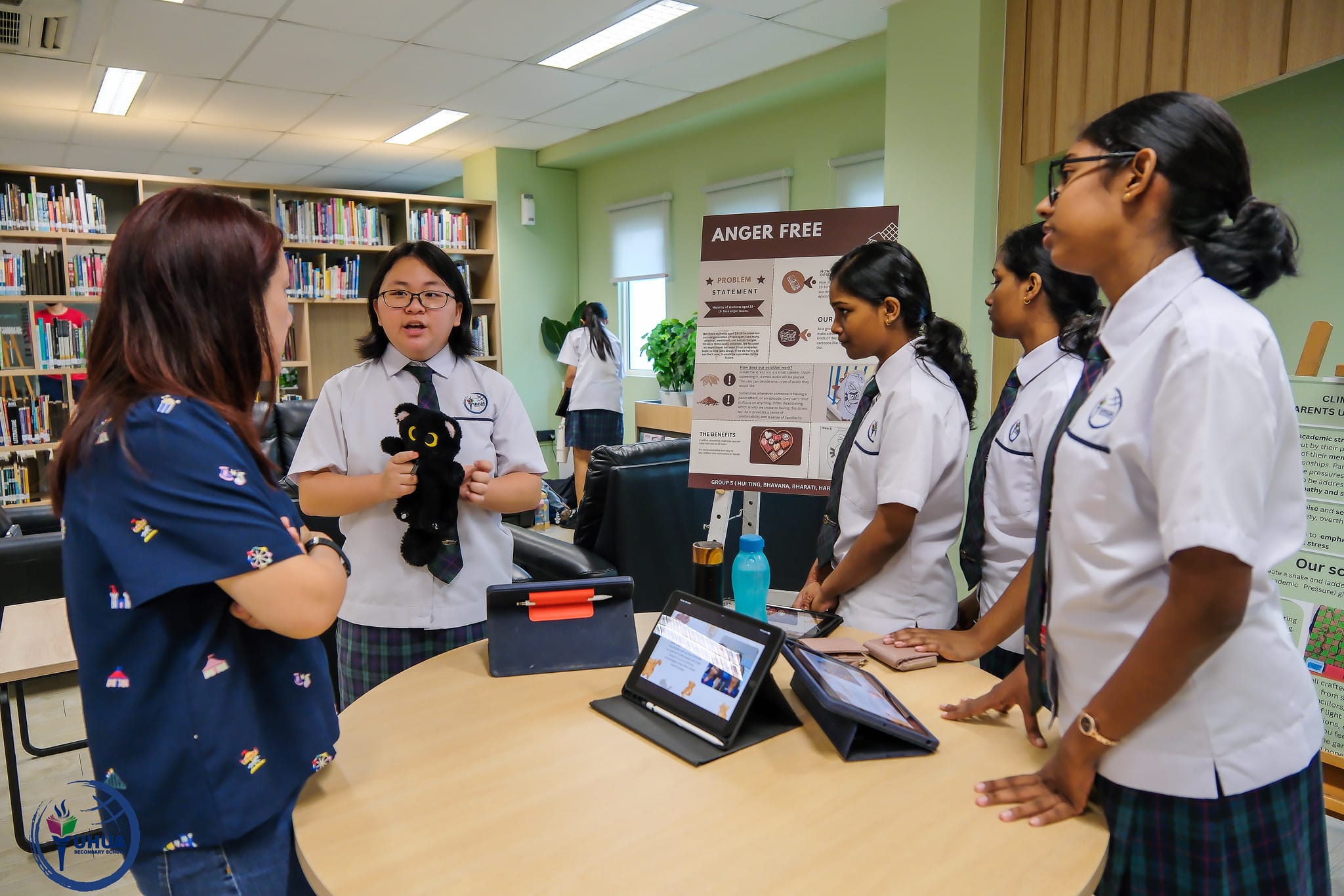 Students in school uniforms present an "Anger Free" project poster to a teacher in a school library.