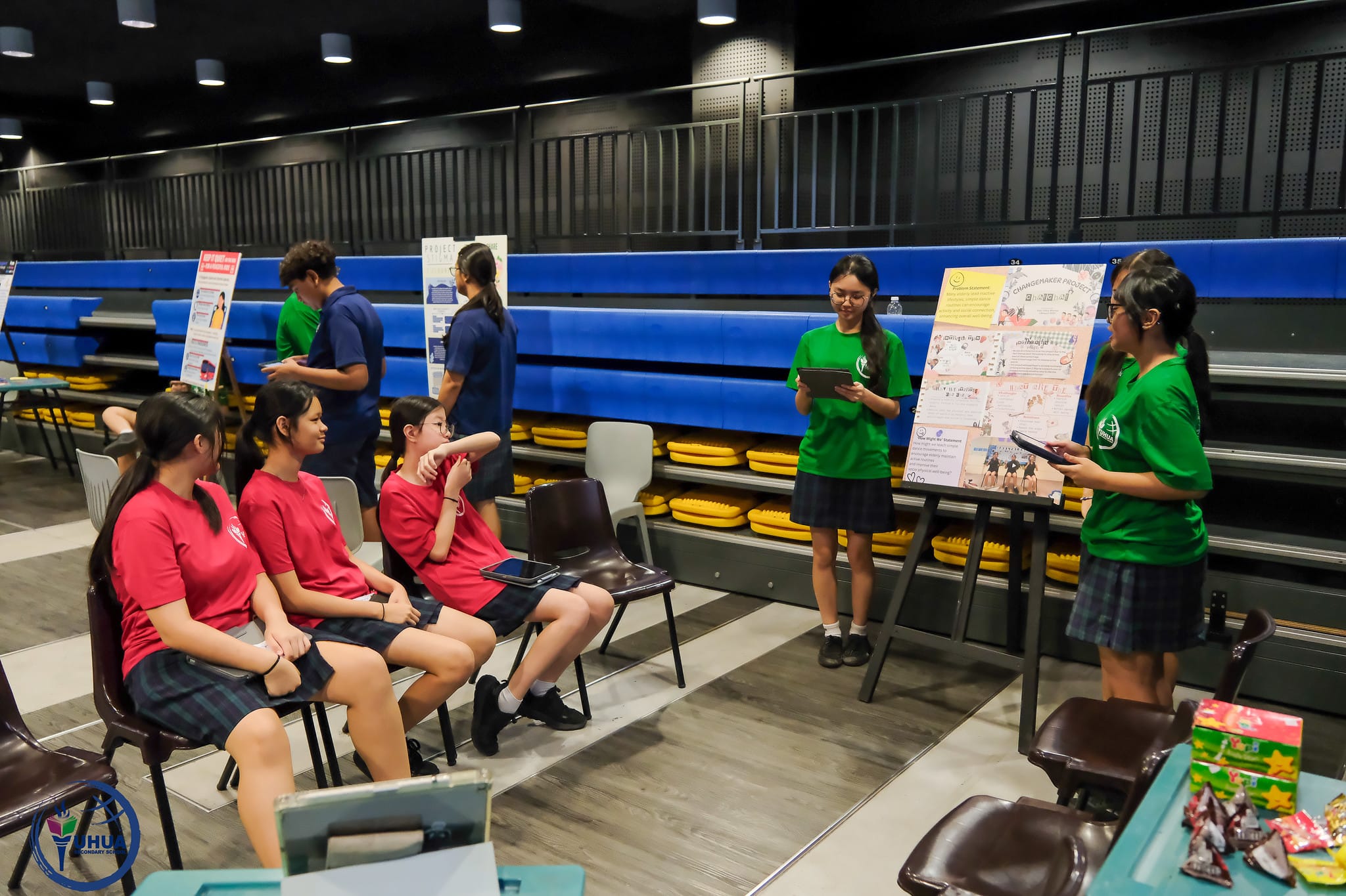 Students in red and green uniforms present project boards to seated peers in a school hall with blue bleachers.