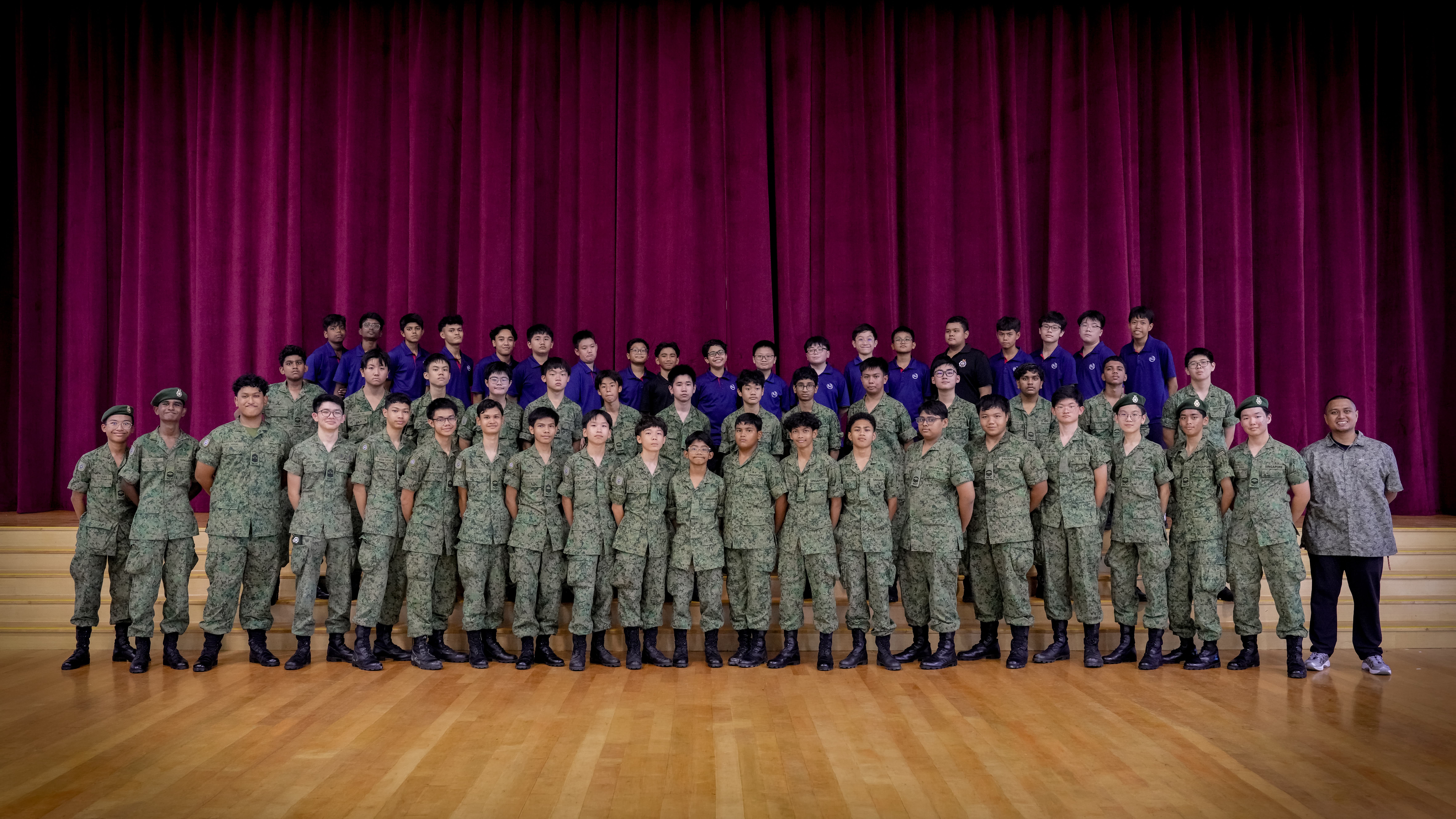 Large group photo of young men in camouflage uniforms and blue shirts on a stage with maroon curtains.