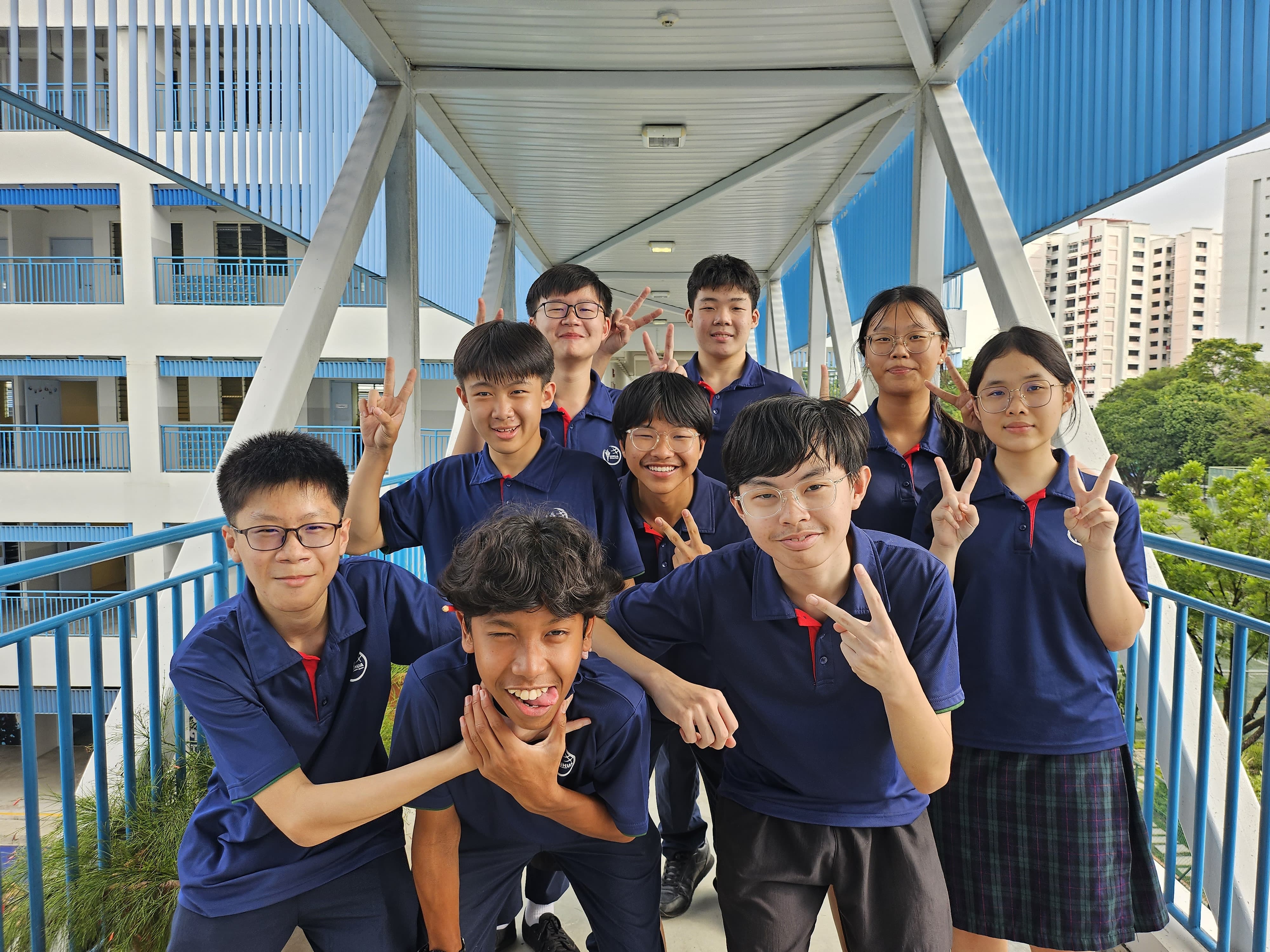 Ten students in navy blue uniforms pose with peace signs on a school walkway with blue railings.