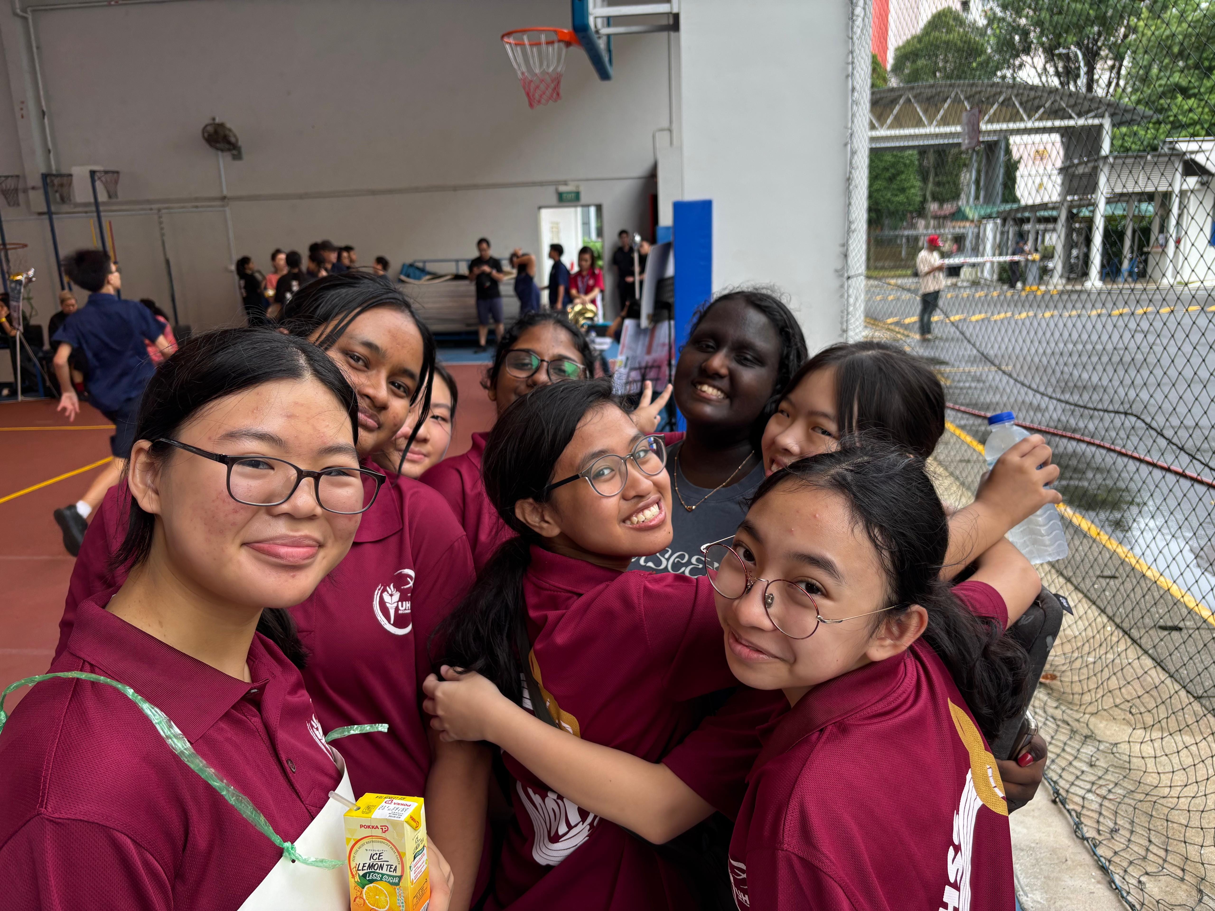 A group of smiling teenage girls in maroon uniforms pose together near a basketball court.