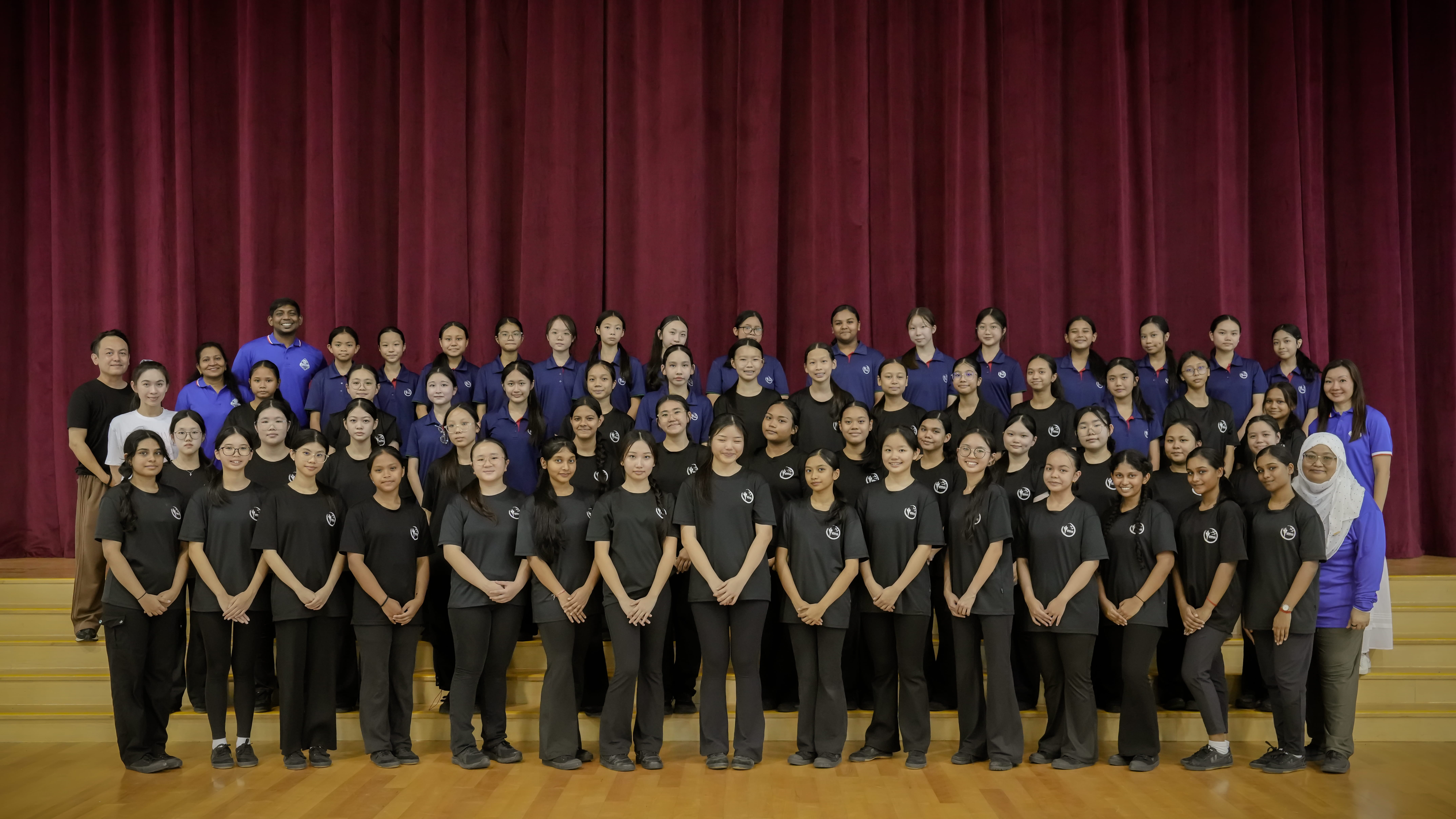 Large group of students and teachers in black and blue shirts posing on stage steps before red curtain