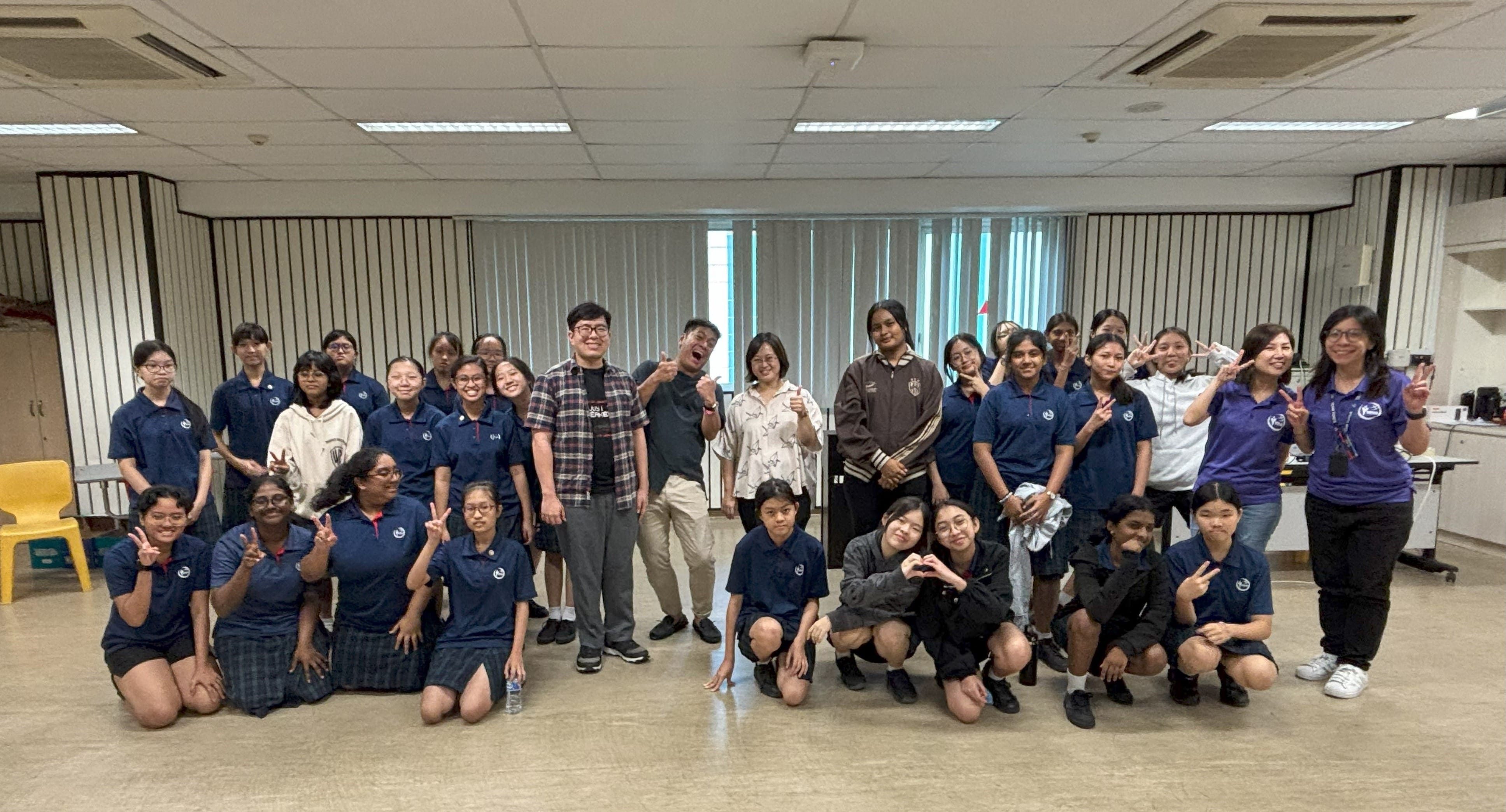 A large group of students in navy uniforms poses with several adults in an indoor room, many smiling and making peace signs.