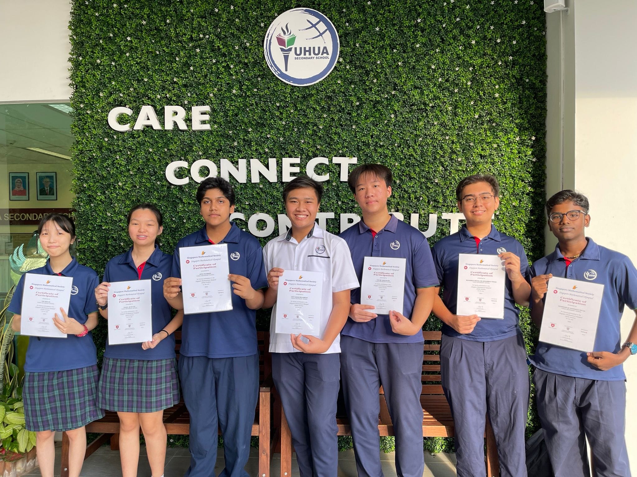 Seven students in blue uniforms hold certificates in front of a green wall with the Yuhua Secondary School logo.