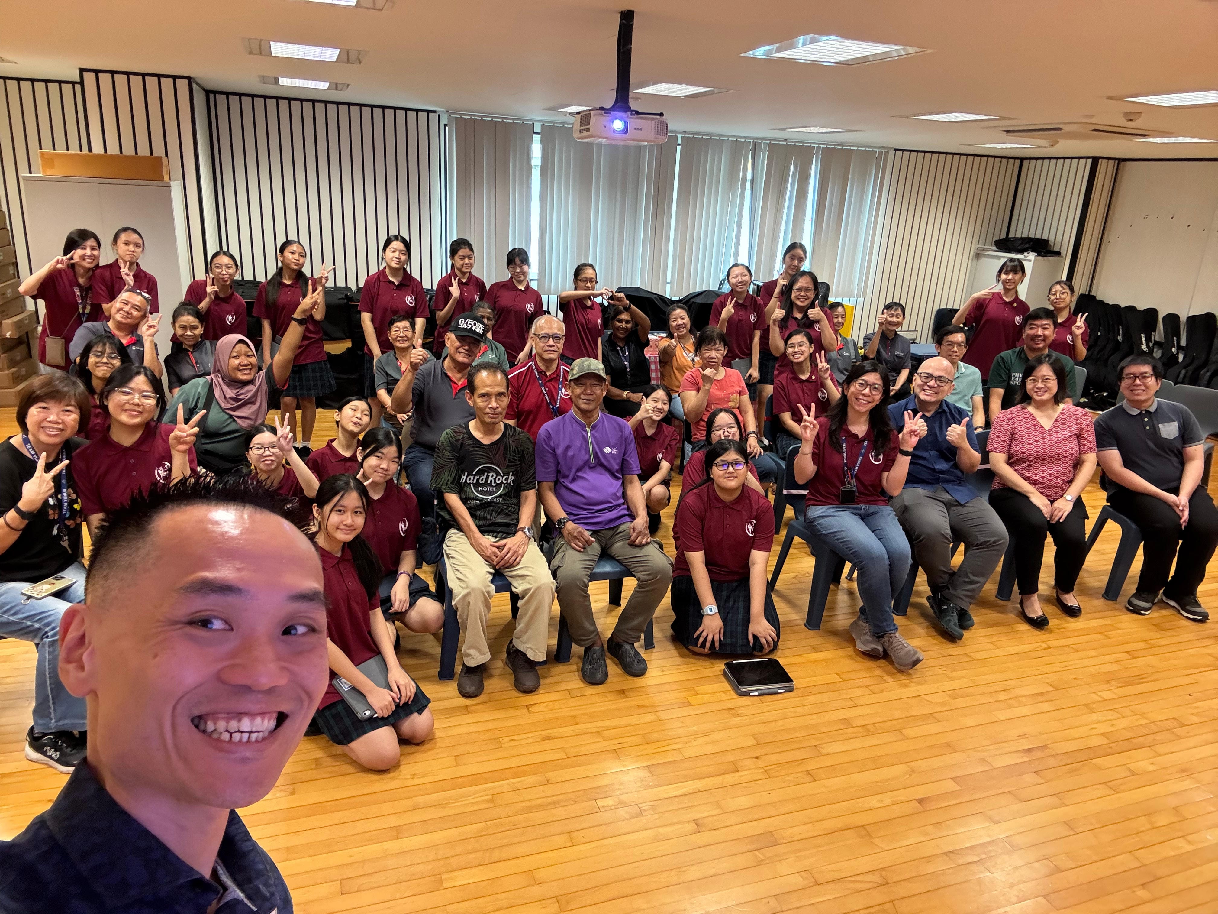 Large group of people in maroon shirts posing with peace signs in an indoor hall, with a selfie taken in the foreground.