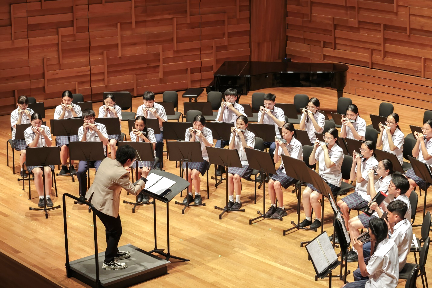 A conductor leads a large group of uniformed students playing harmonicas on a wooden concert hall stage.