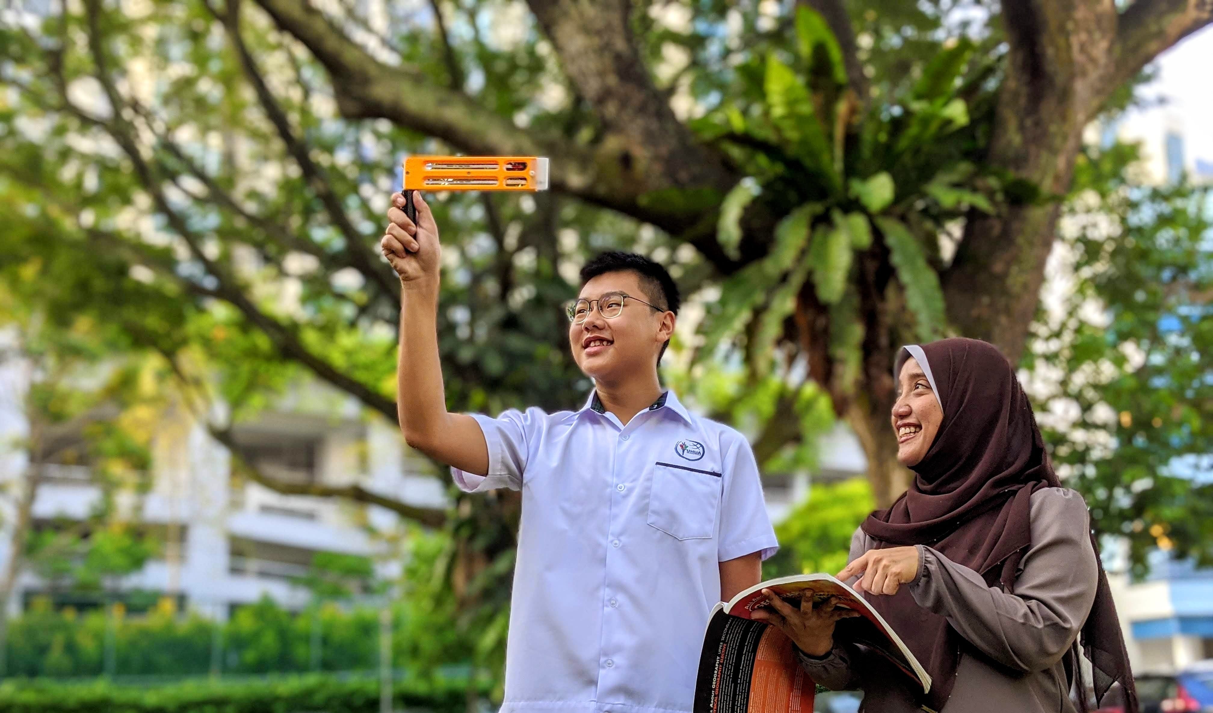 Two smiling students outdoors, one holding an orange tool up, the other holding an open book, trees in background.