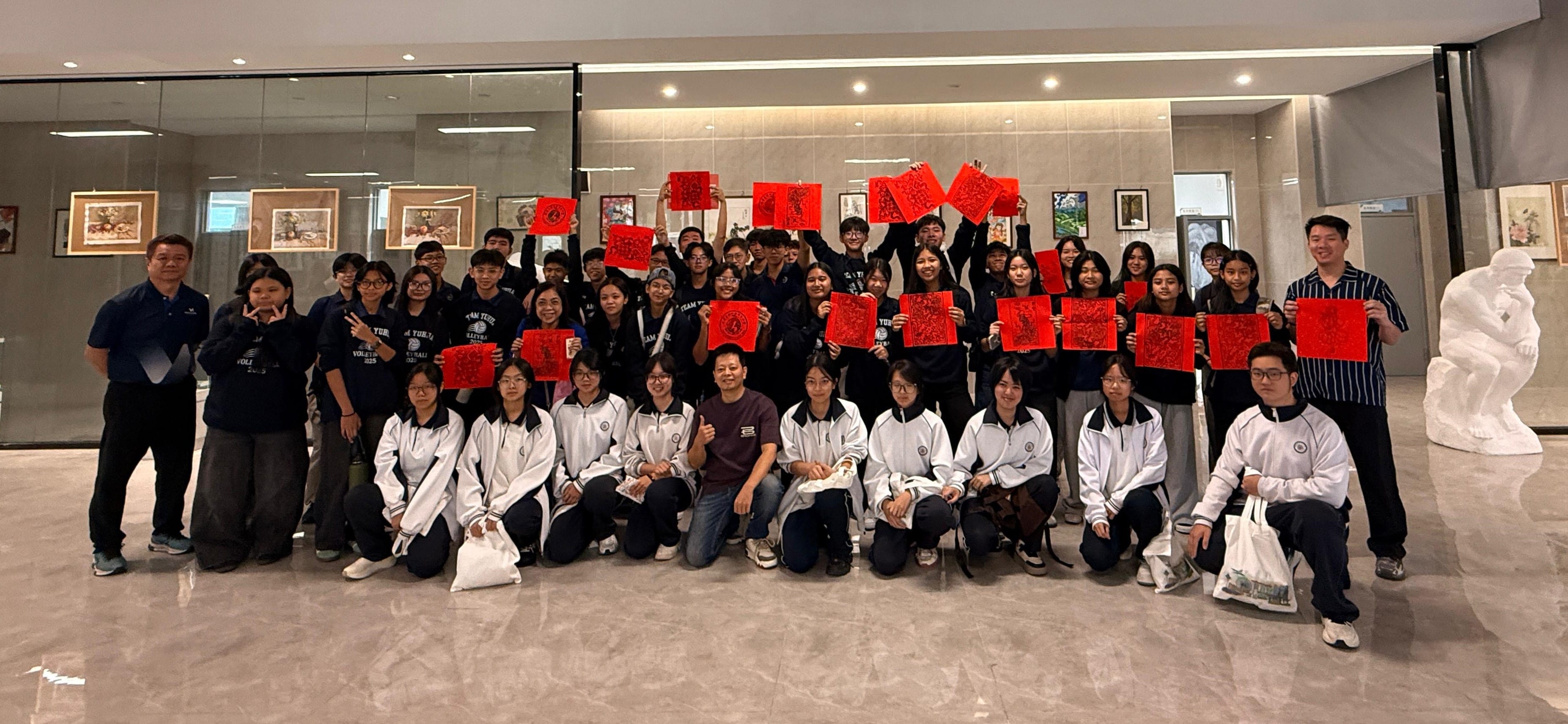 A large group of students and adults pose indoors holding red paper-cut artworks, with framed paintings on the wall behind them.