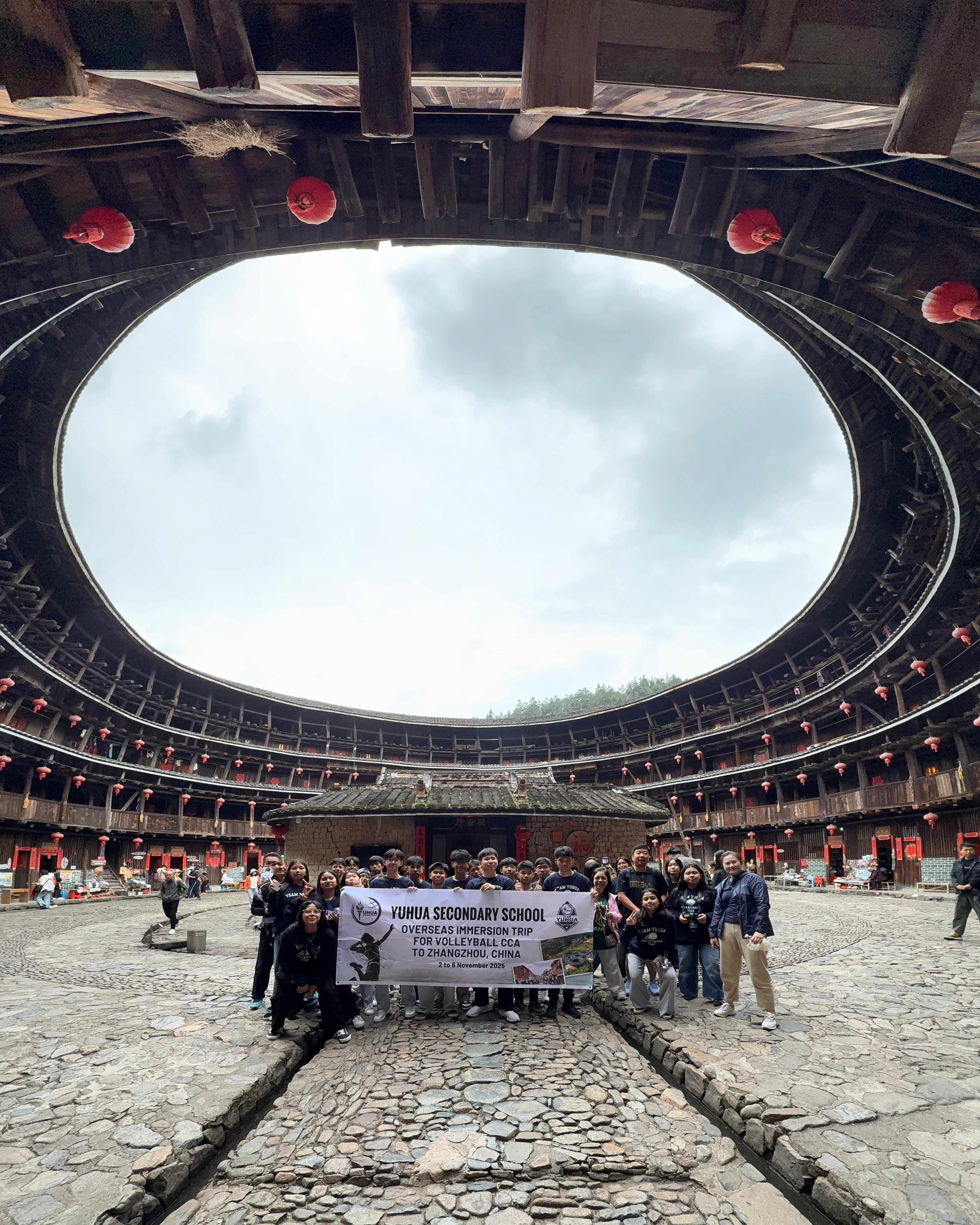 Group of students holding a Yuhua Secondary School banner inside a circular Fujian tulou building with red lanterns.