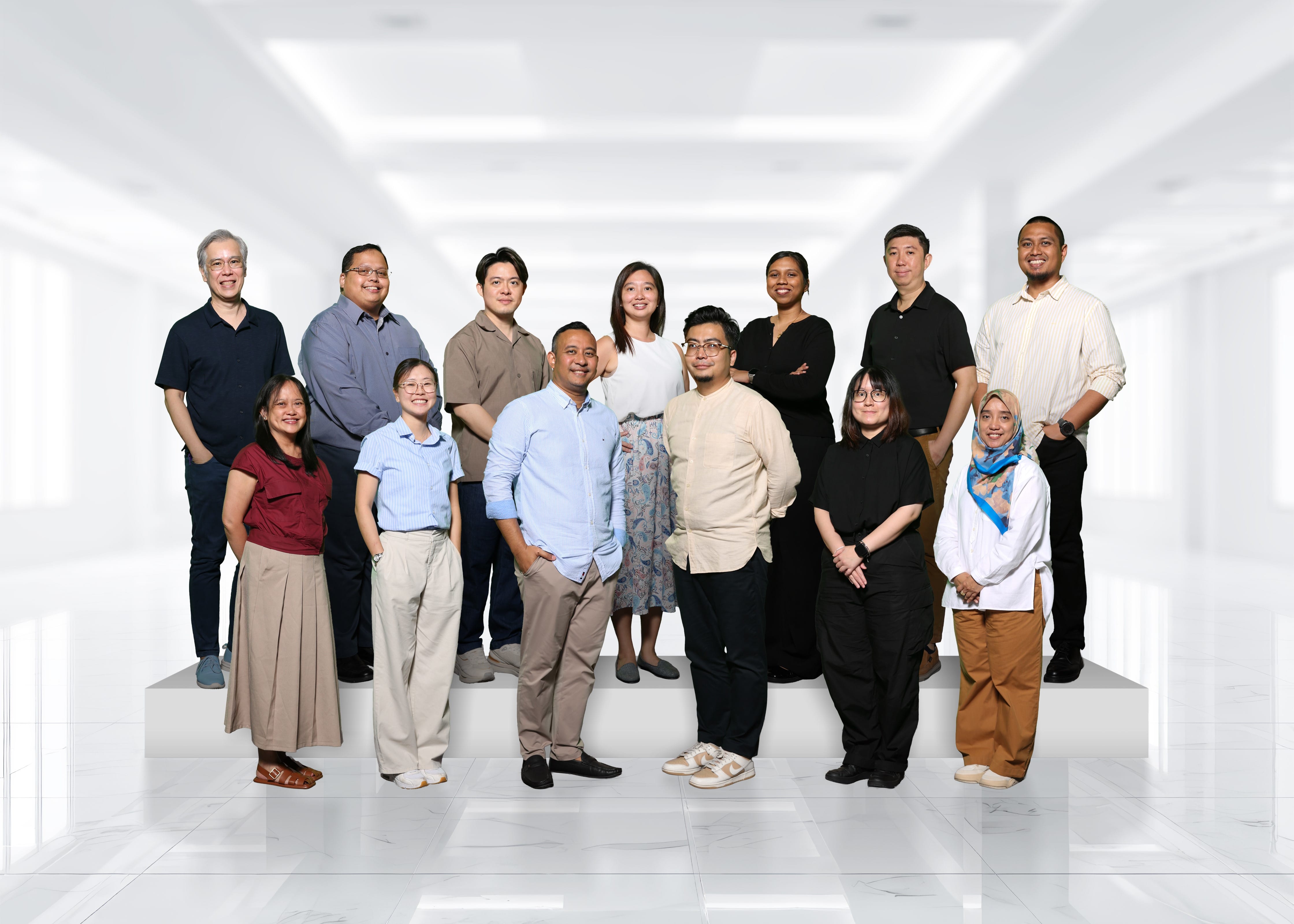 Thirteen people posing together in two rows against a bright white indoor background, smiling at the camera.