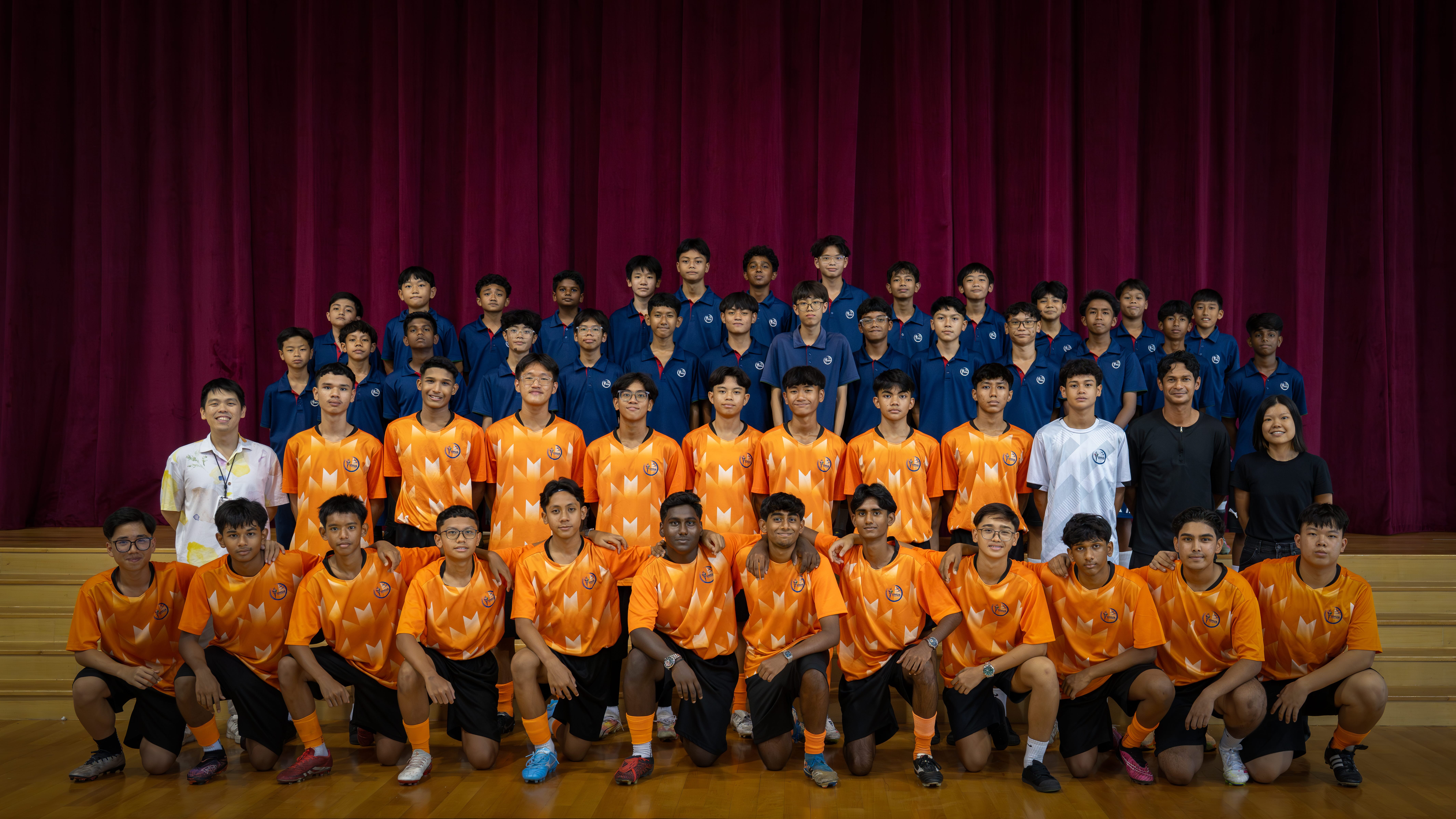 Group photo of boys in orange sports jerseys and navy uniforms posed in rows before maroon curtains