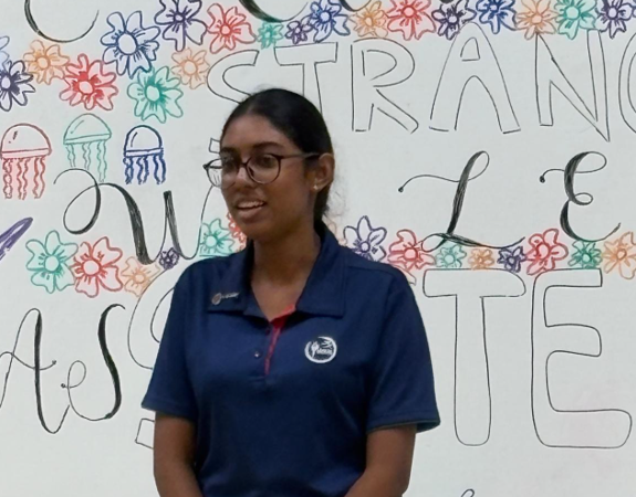 Young woman with glasses in a navy polo shirt standing before a colorful hand-drawn mural with flowers and text.