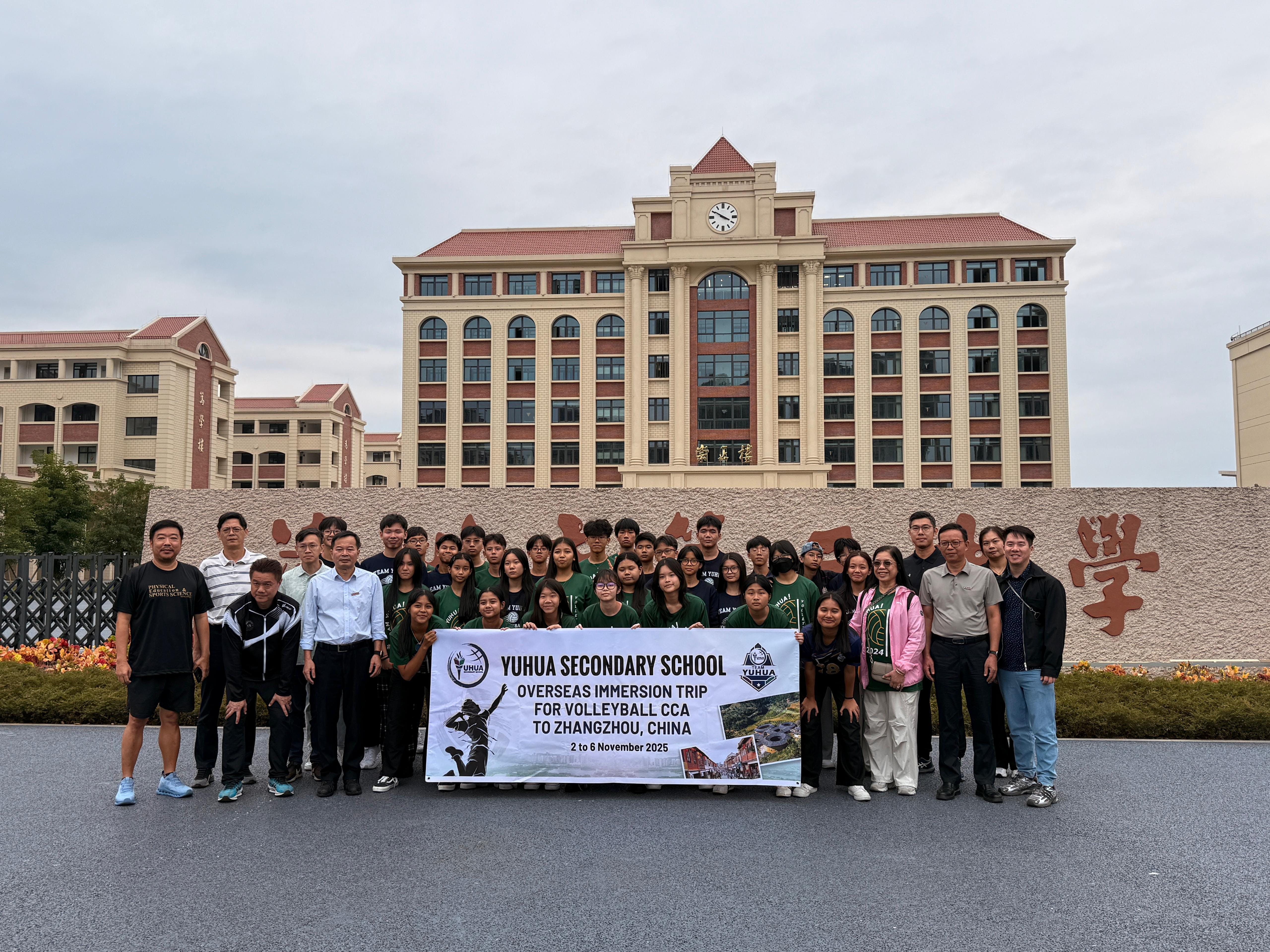 Group of students and adults holding a Yuhua Secondary School volleyball trip banner in front of a large building in China.