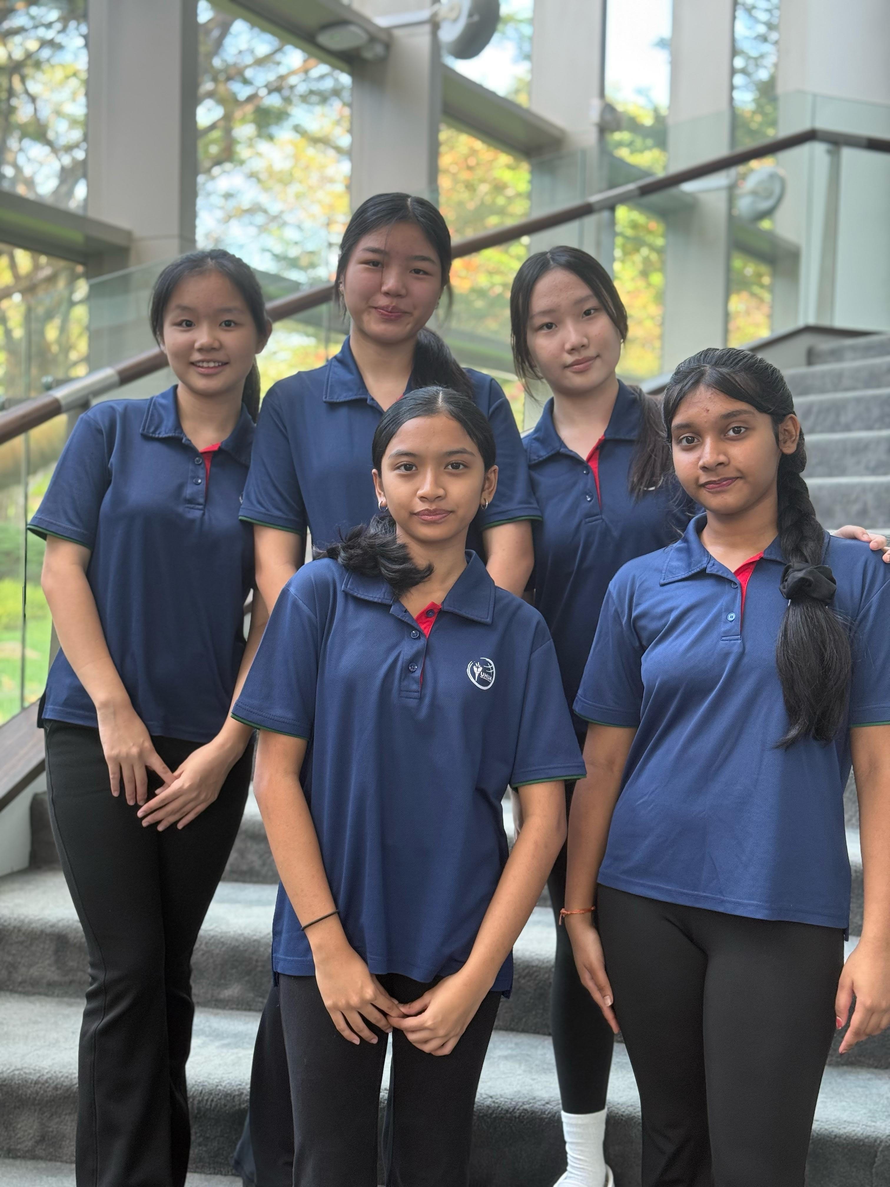 Five young women in matching blue polo shirts pose together on an indoor staircase with glass windows behind them.