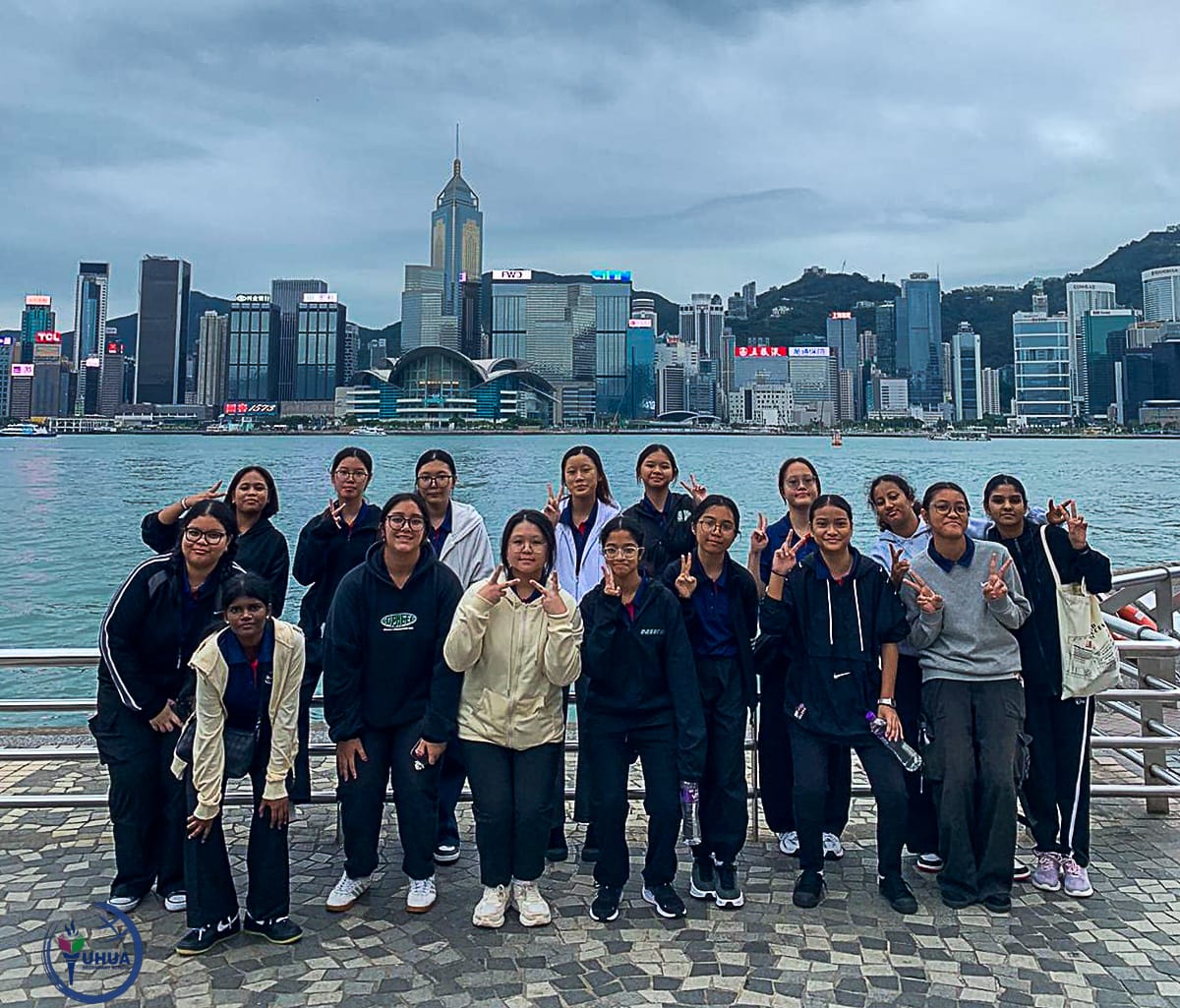 A group of young women posing with peace signs on a waterfront promenade with Hong Kong skyline behind them.