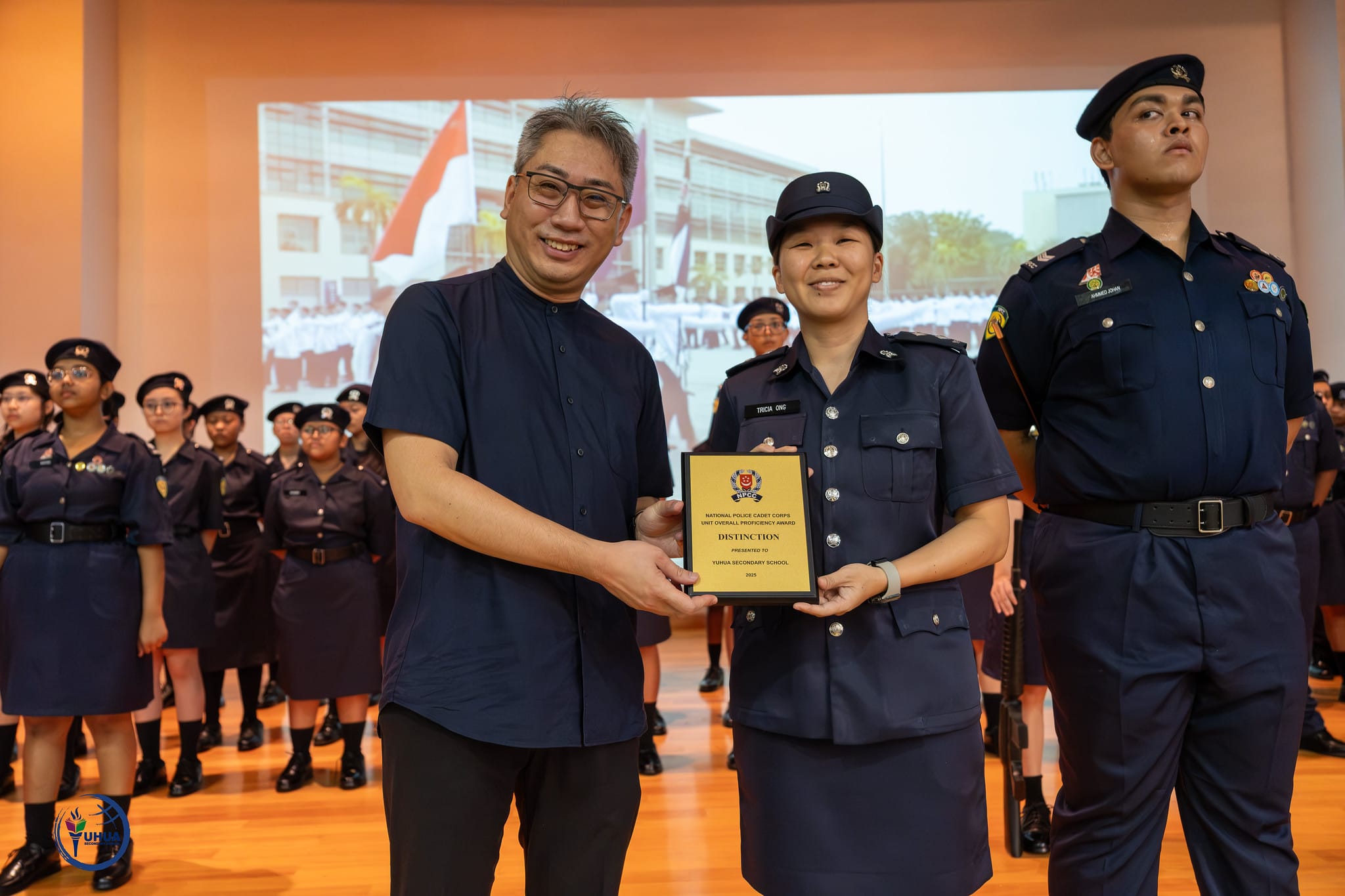 A man and a uniformed cadet hold a Distinction plaque from the National Police Cadet Corps at a ceremony.