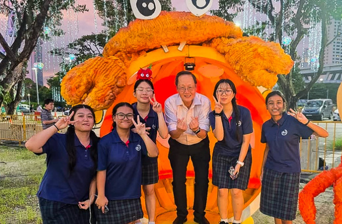 Six people pose in front of a large orange crab sculpture outdoors, five in school uniforms making peace signs.