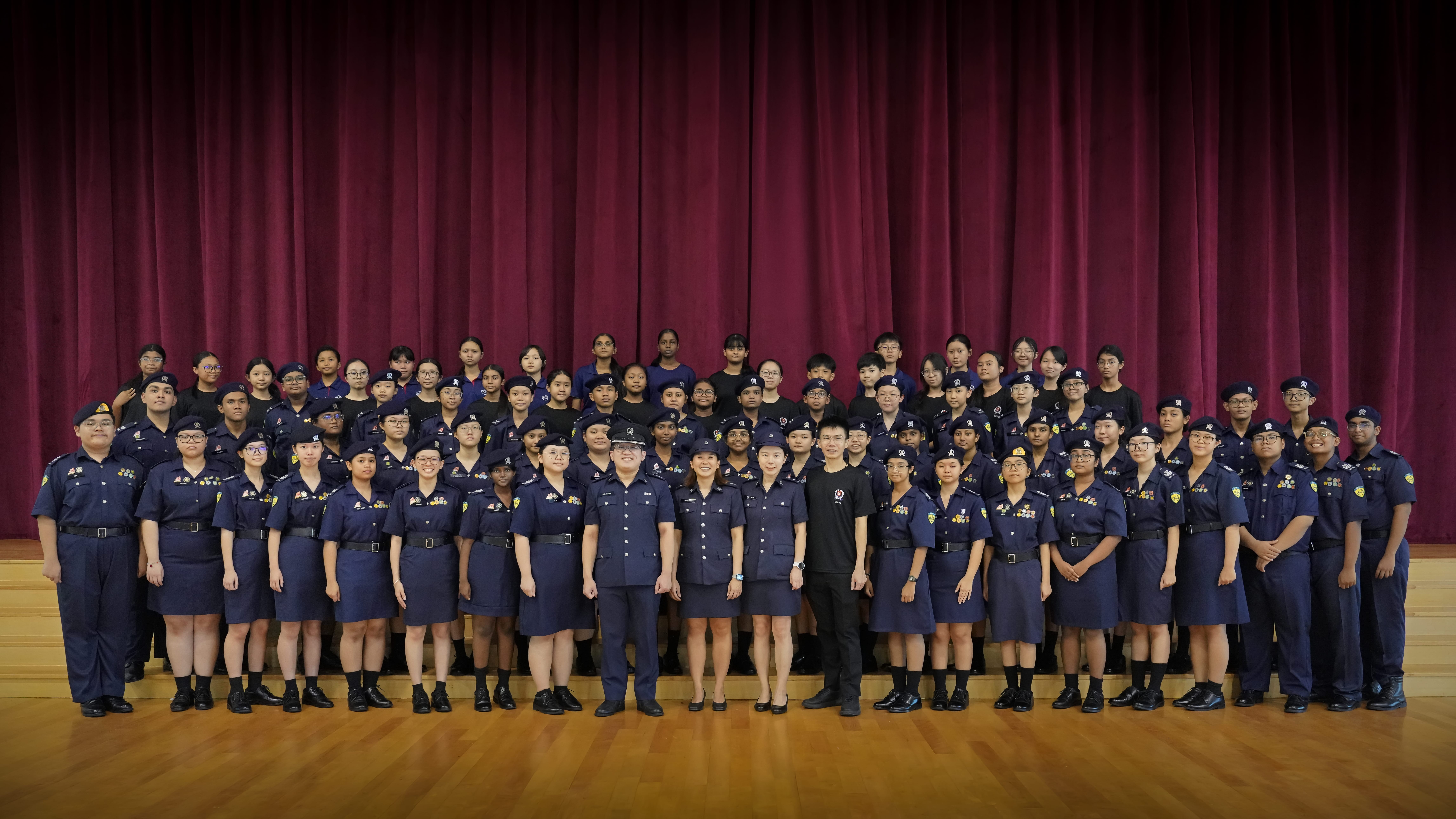 Large group in navy uniforms posing on a stage, with a red curtain backdrop and wooden floor.