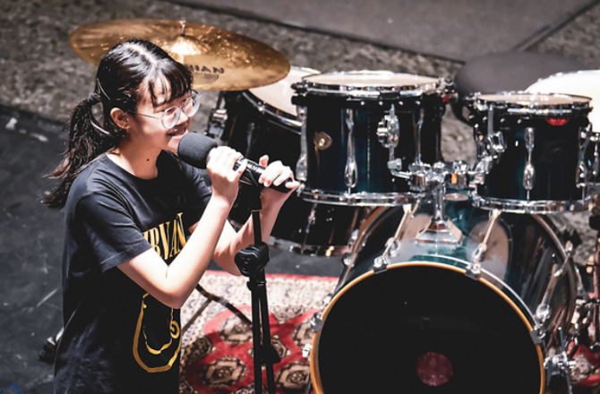 Young woman with glasses and ponytail singing into a microphone on stage with a drum kit behind her.