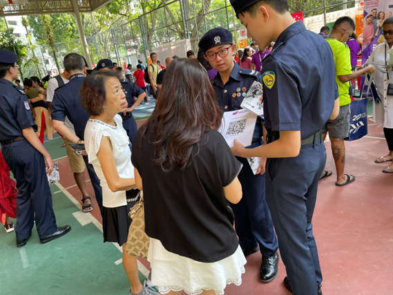 Two uniformed security officers speak with two women at a crowded outdoor event on a red court.