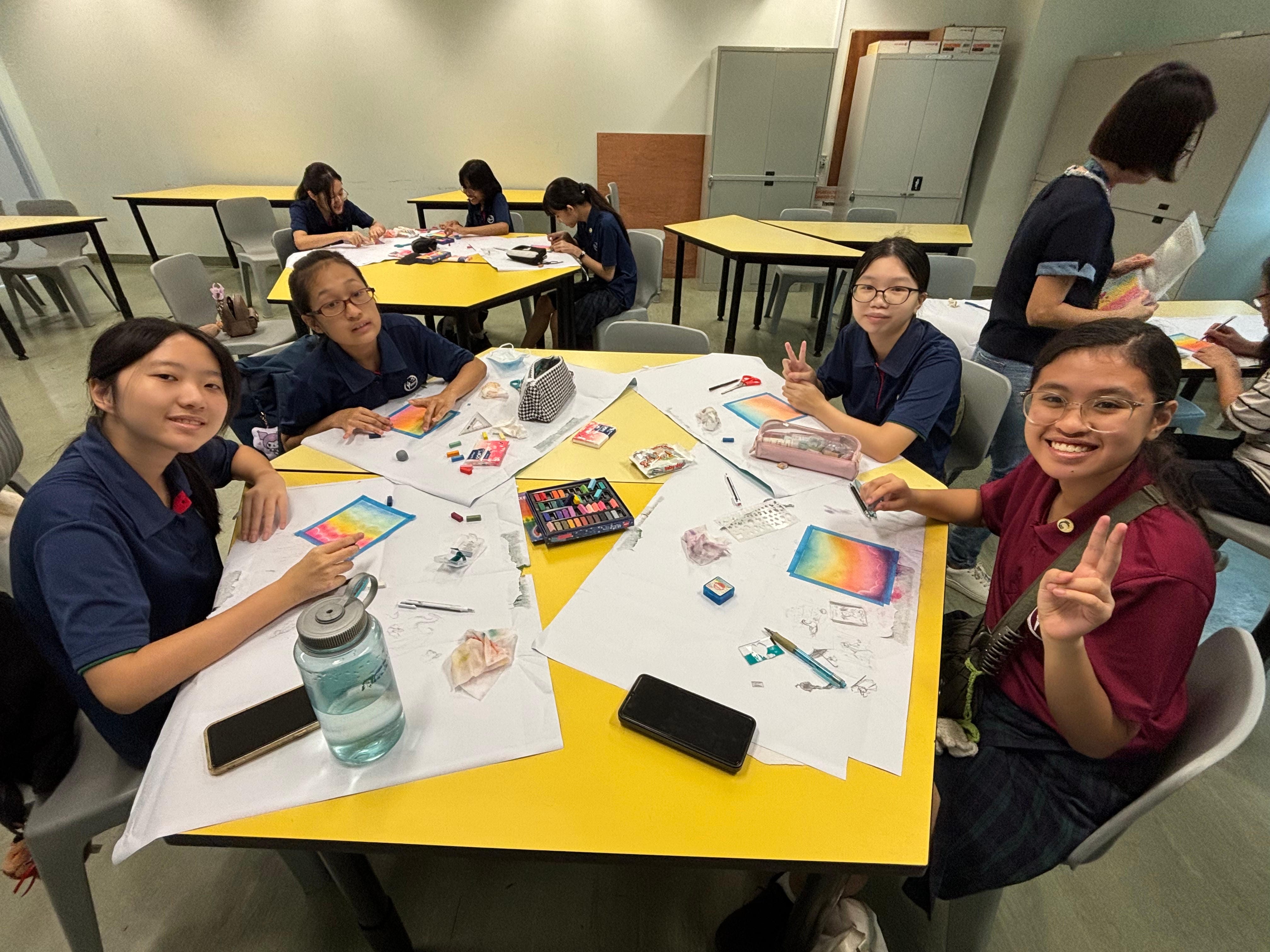 Students in blue uniforms sit at yellow tables doing pastel artwork, smiling and making peace signs at the camera.