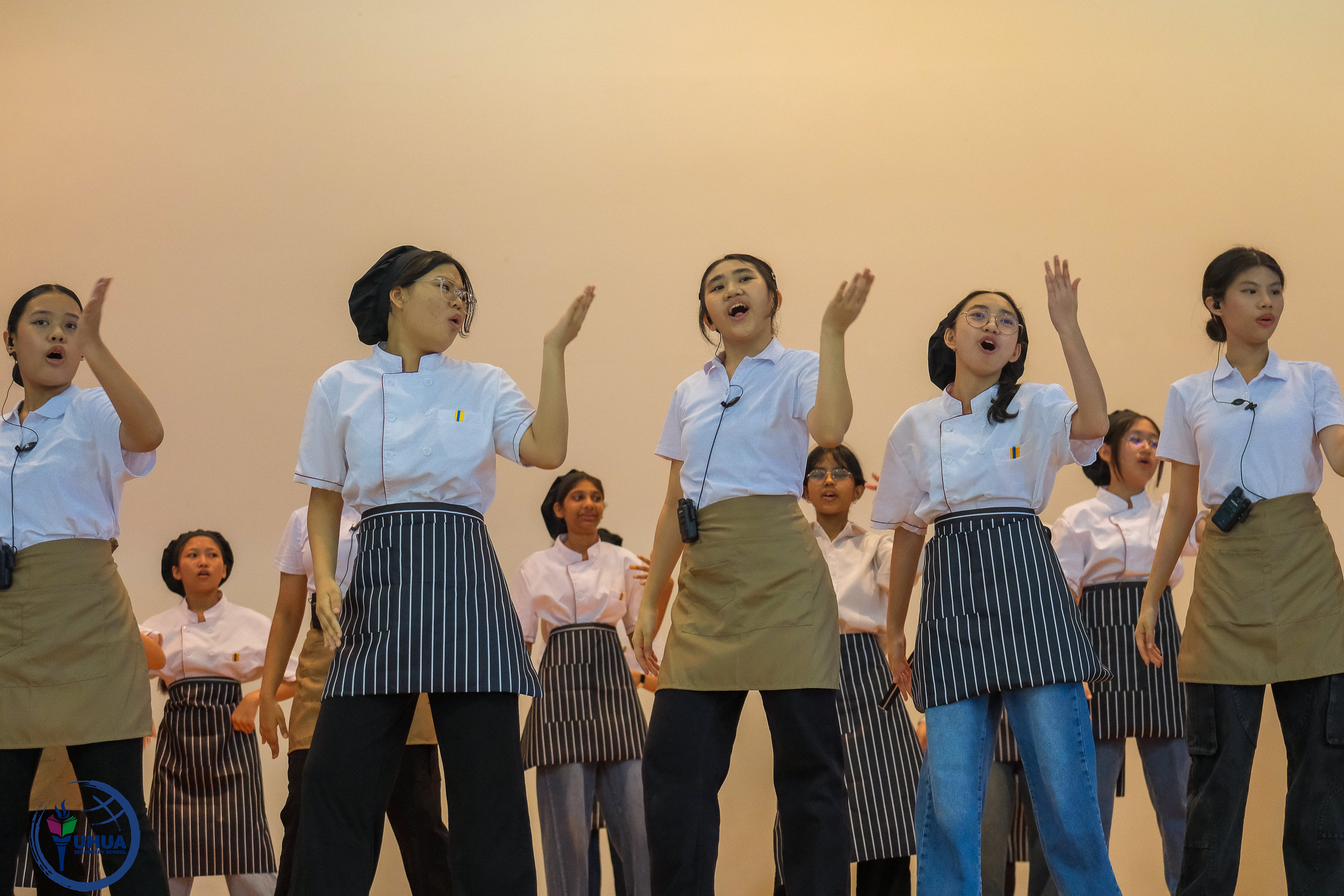 Group of students in white uniforms and aprons sing on stage, hands raised, wearing headset mics.