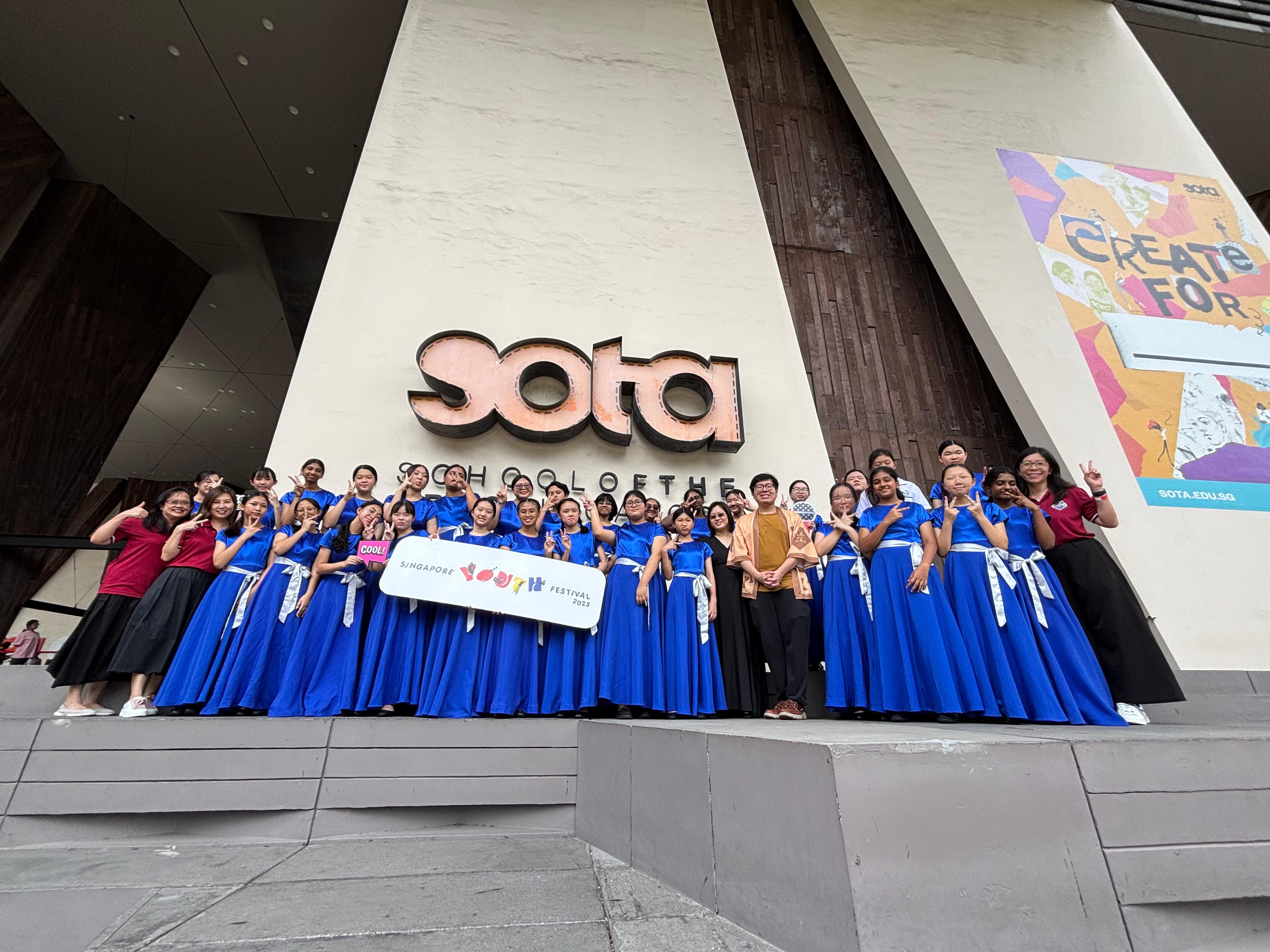 A group of performers in blue gowns pose with a Singapore Youth Festival 2023 sign outside the SOTA building.